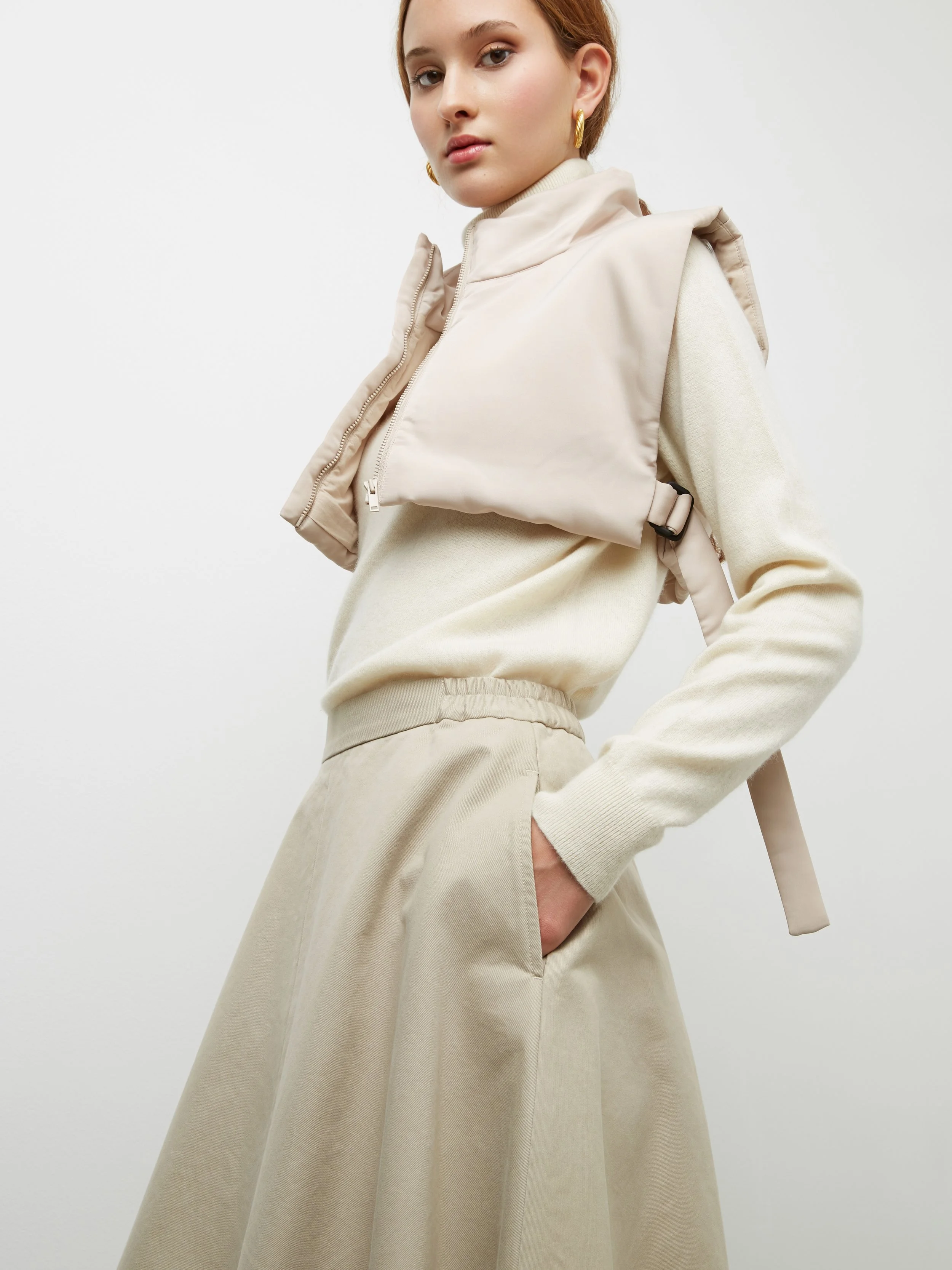 Donna con giacca in pile crema, pantaloni beige a vita elasticizzata, portando una giacca beige sulle spalle, con orecchini dorati e capelli raccolti.