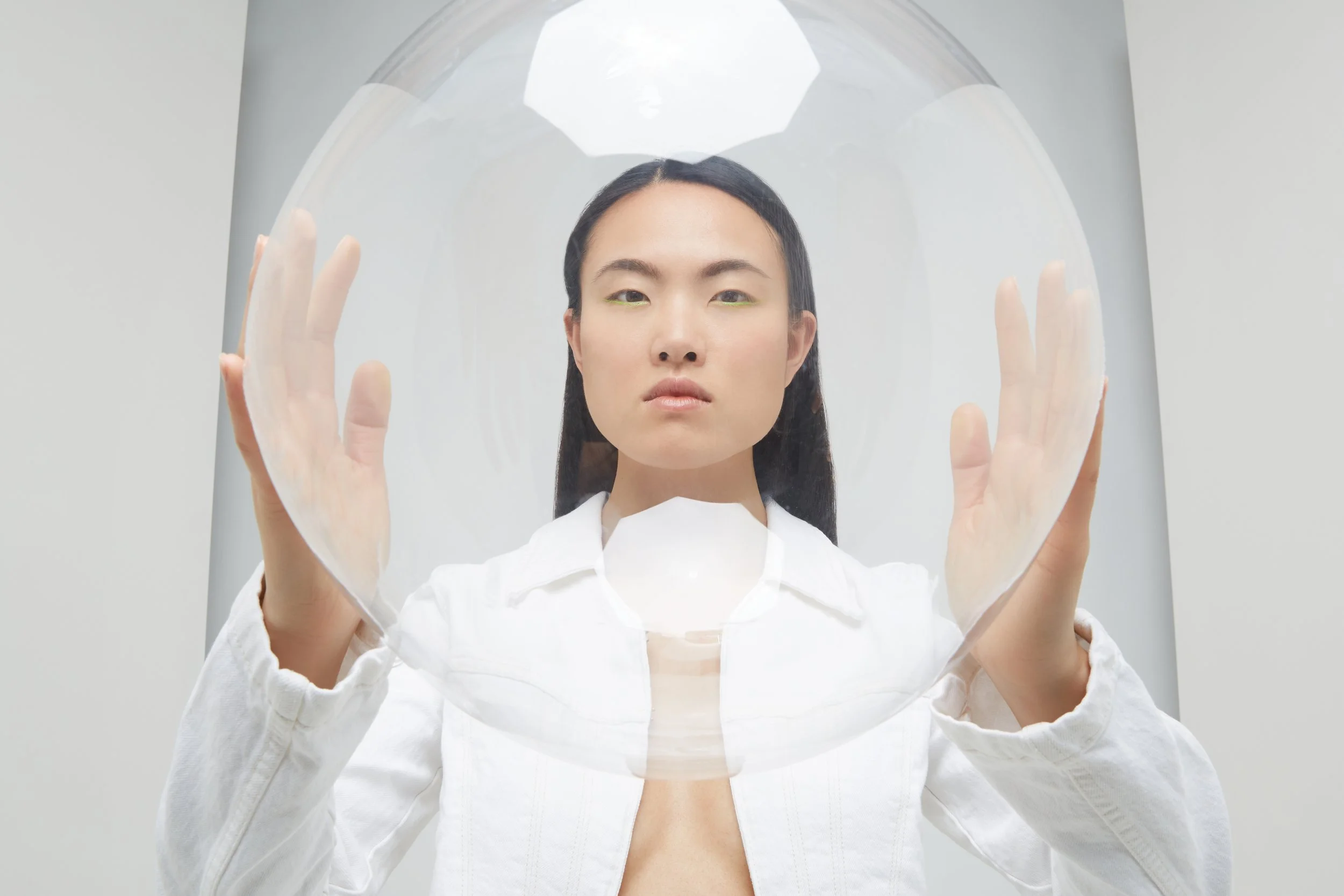 Una donna asiatica con capelli lisci e trucco naturale che si trova dietro una lastra di vetro trasparente e tiene le mani contro di essa.