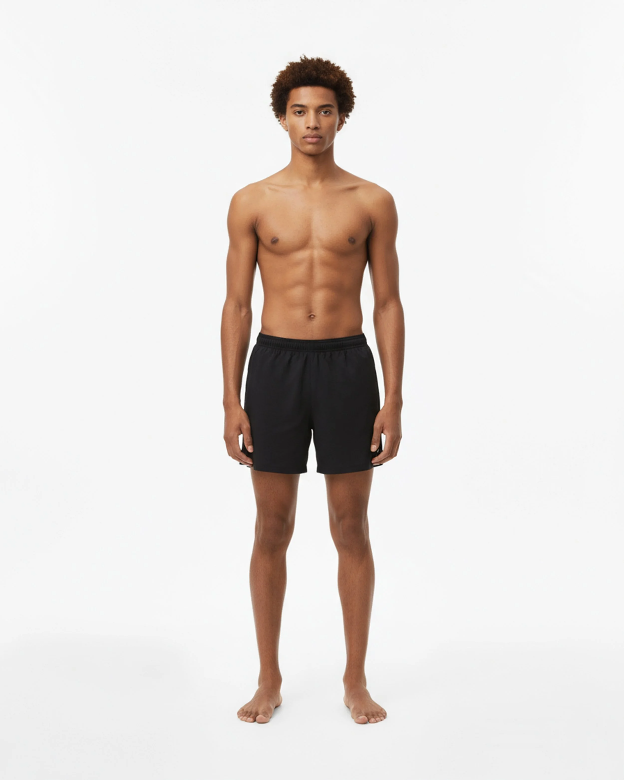 Un giovane uomo nudo con pantaloncini neri si trova davanti a uno sfondo bianco.