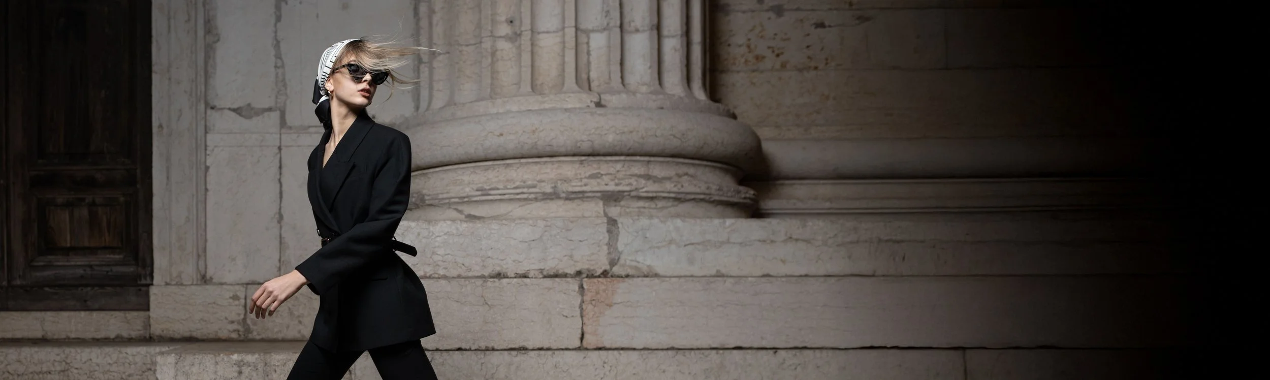 Una donna elegante con occhiali da sole e cappello cammina davanti a colonne di marmo, indossando un completo nero raffinato.