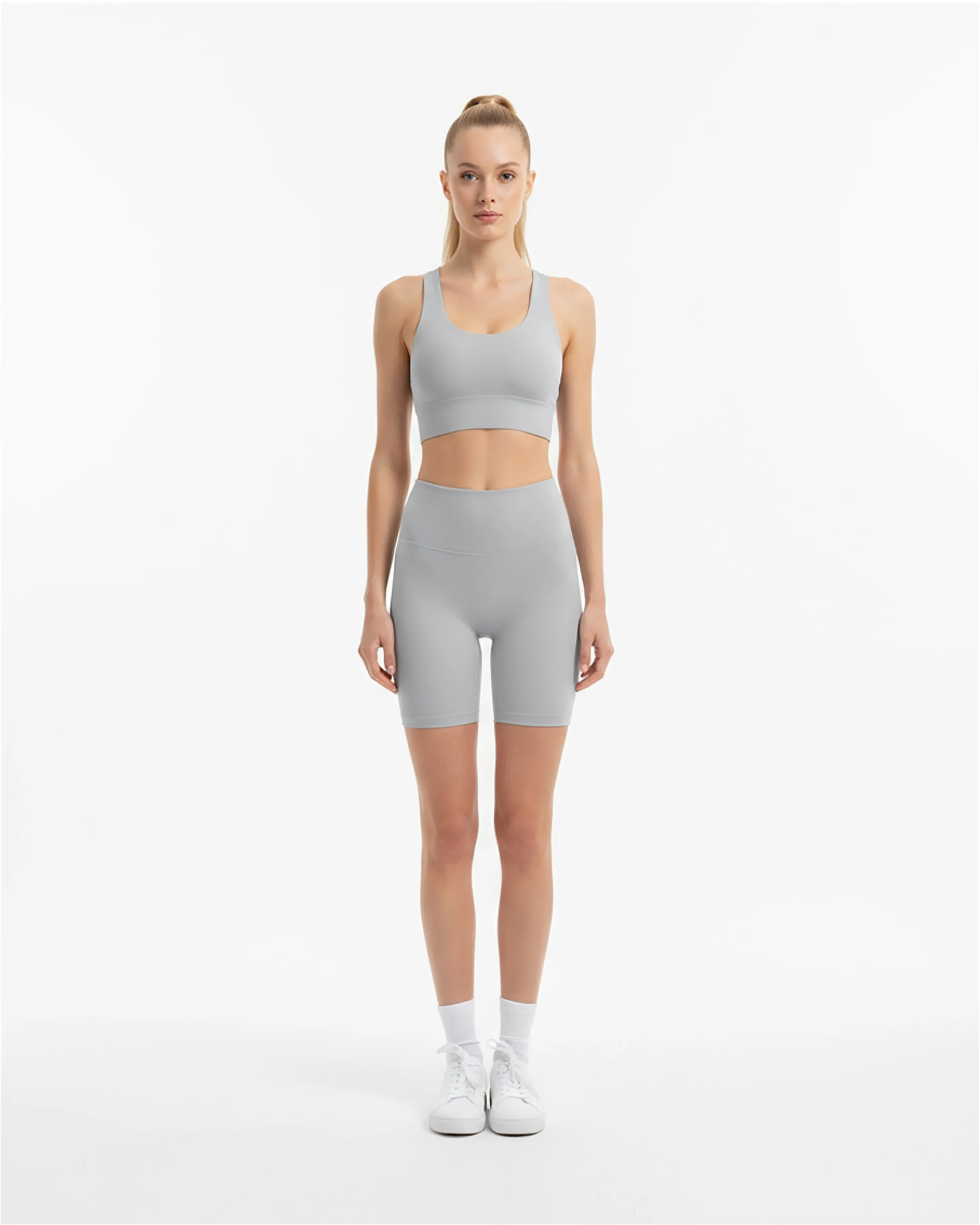 Donna che indossa abbigliamento sportivo grigio, composta da reggiseno, shorts e scarpe da ginnastica, in piedi su sfondo bianco.