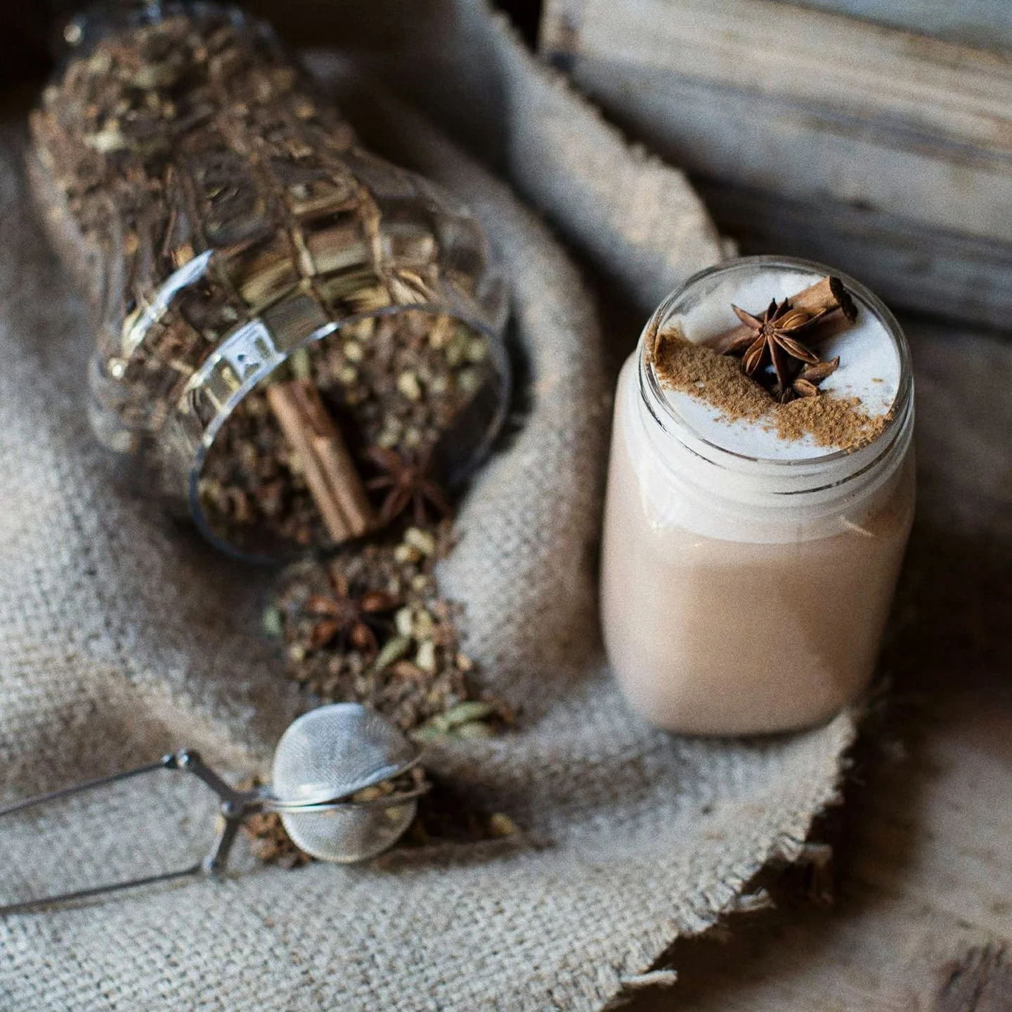 Bevanda in un barattolo di vetro con panna, cannella e anice stellato sopra, circondata da spezie come chiodi di garofano, cannella in stecco e pepe, con un colino e un barattolo di spezie sul tessuto di juta.