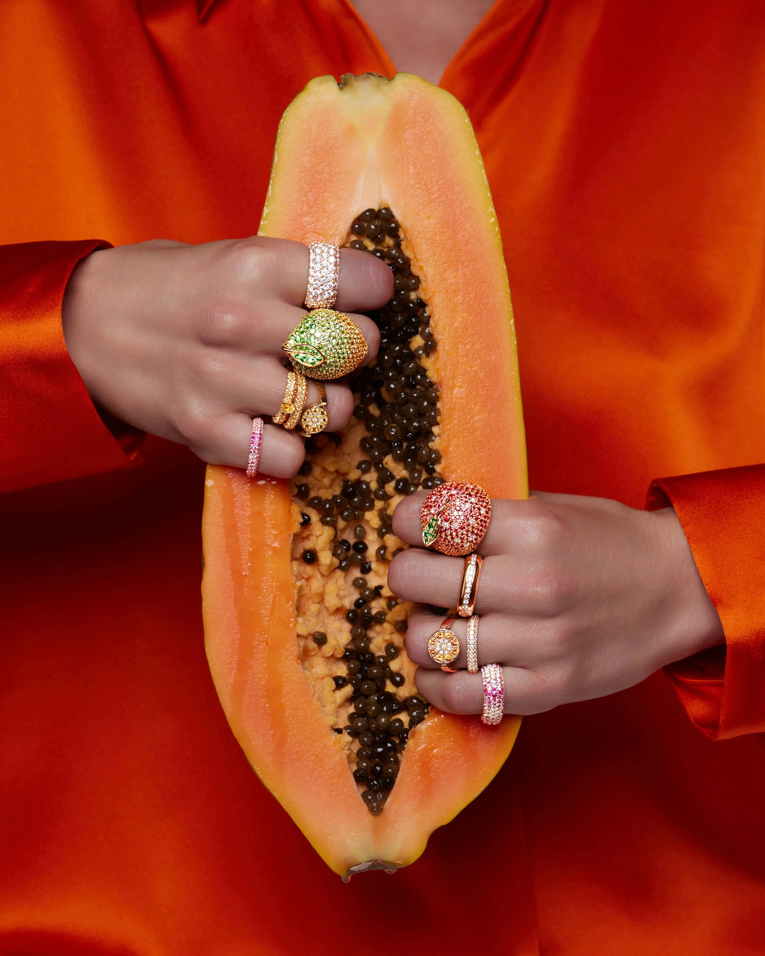 Due mani indossano vari anelli decorativi, una tiene mezza papaya aperta, con semi neri all'interno, su sfondo arancione e abiti arancioni.