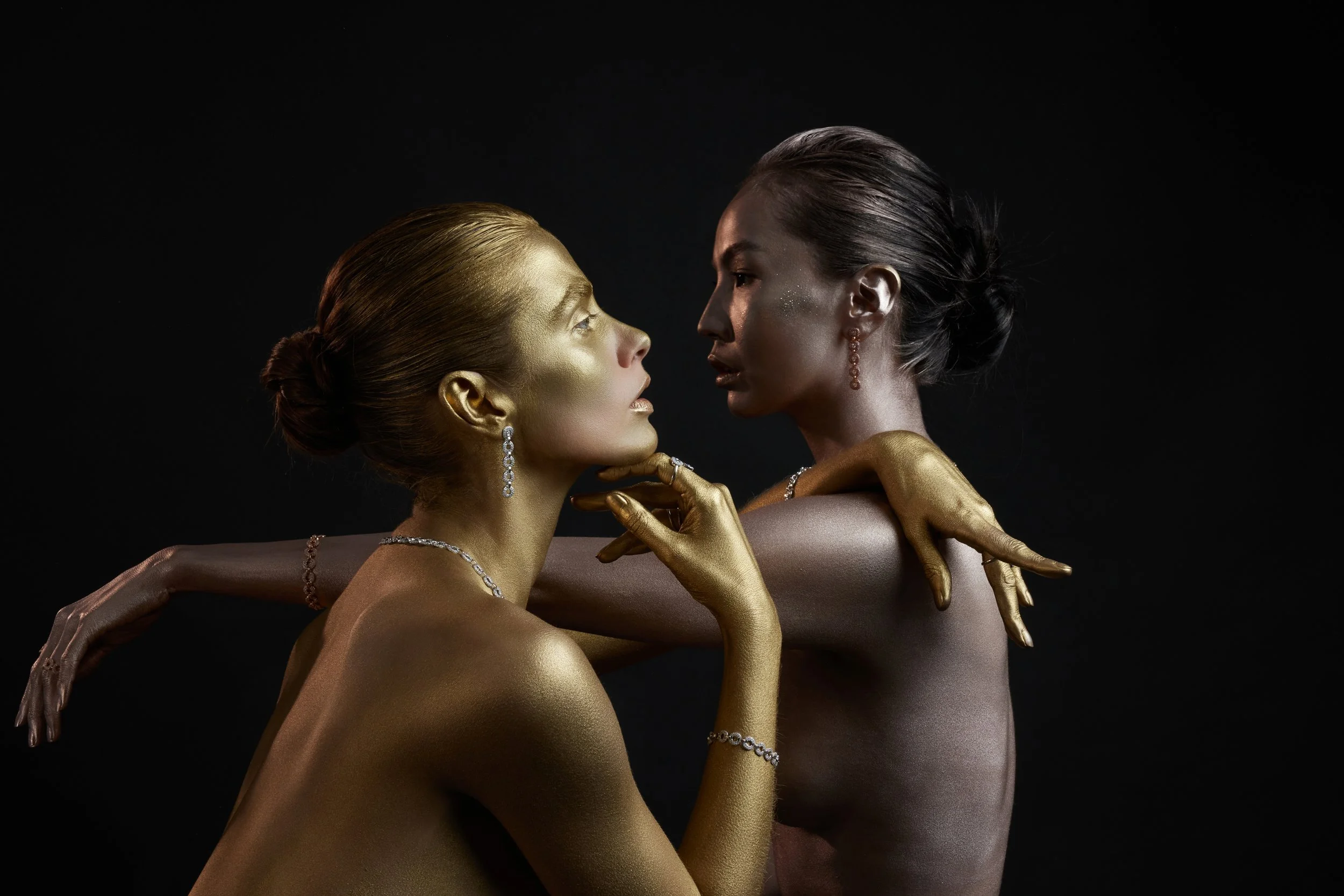 Due donne senza abiti, dipinte di oro, si incontrano in un'ambientazione scura. Una donna tiene il viso dell'altra tra le dita, entrambe indossano gioielli e hanno i capelli raccolti.