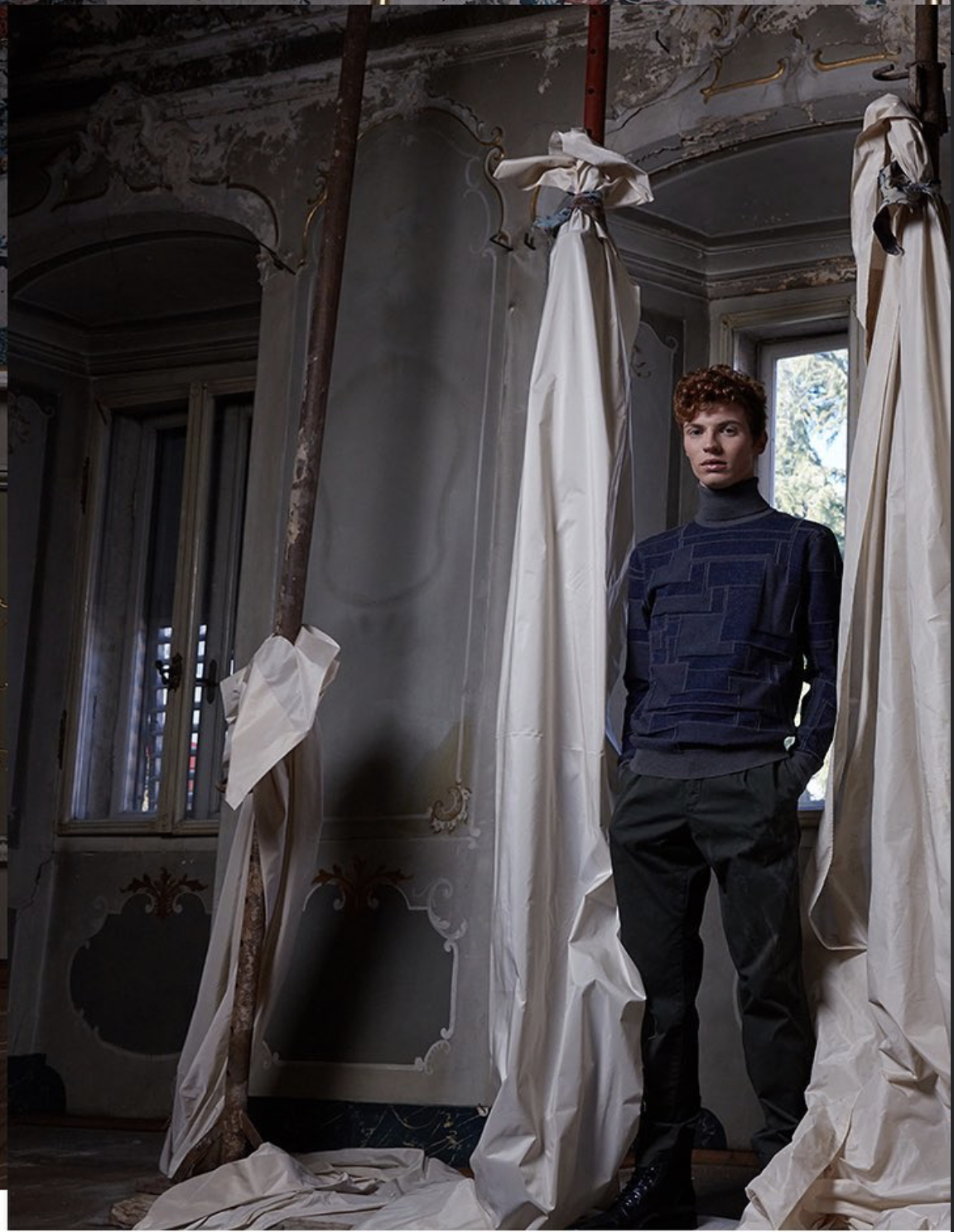 Un uomo giovane con i capelli ricci e indossa un maglione blu e pantaloni neri, si trova in una stanza con pareti decorate e grandi finestre. È circondato da tende di tessuto bianco, alcune delle quali sono sospese o appoggiate a terra.
