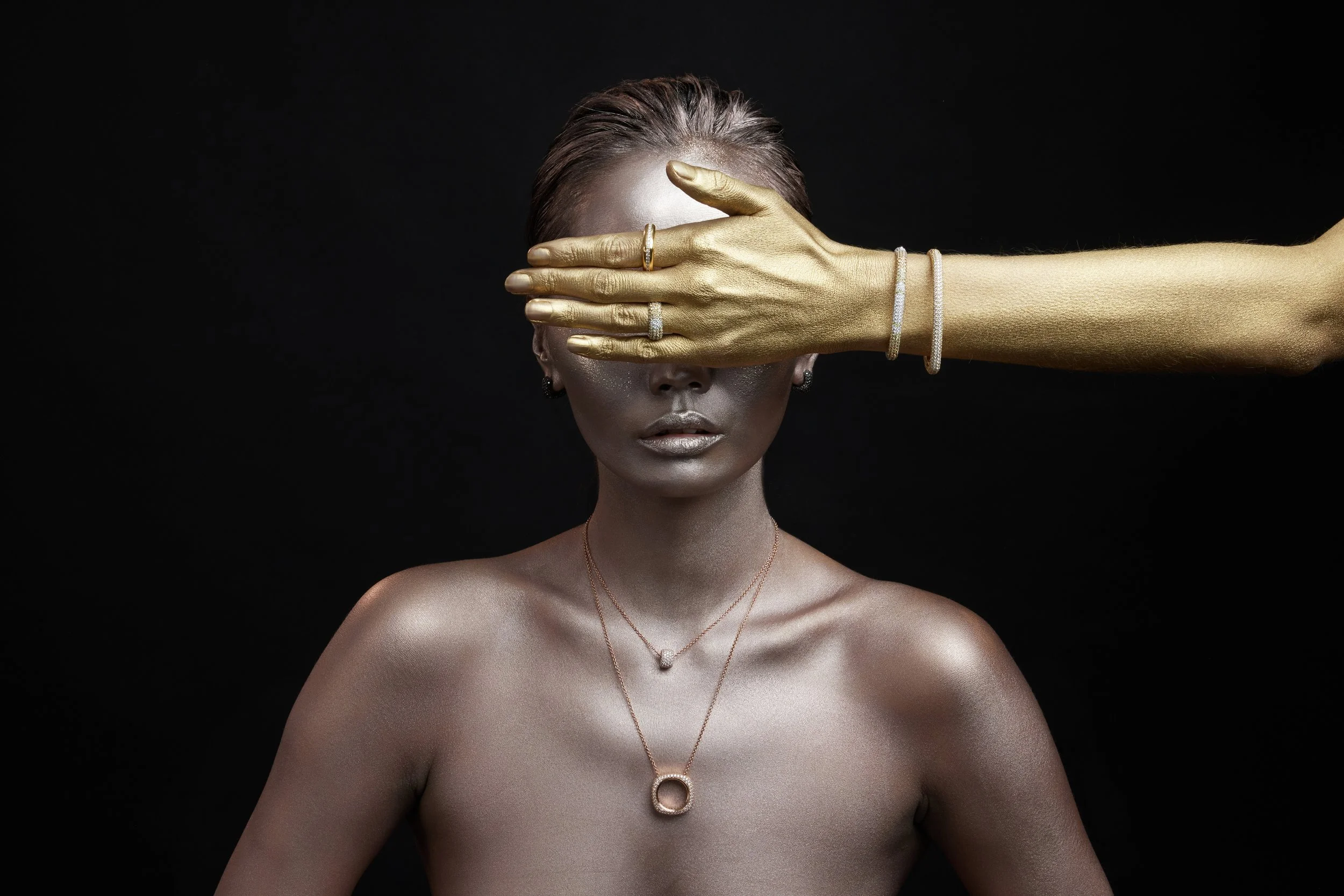 Una donna con la pelle color argento o con trucco lucido, indossa collane e orecchini, e ha i capelli corti. Una mano dorata copre gli occhi della donna, decorata con anelli e braccialetti.