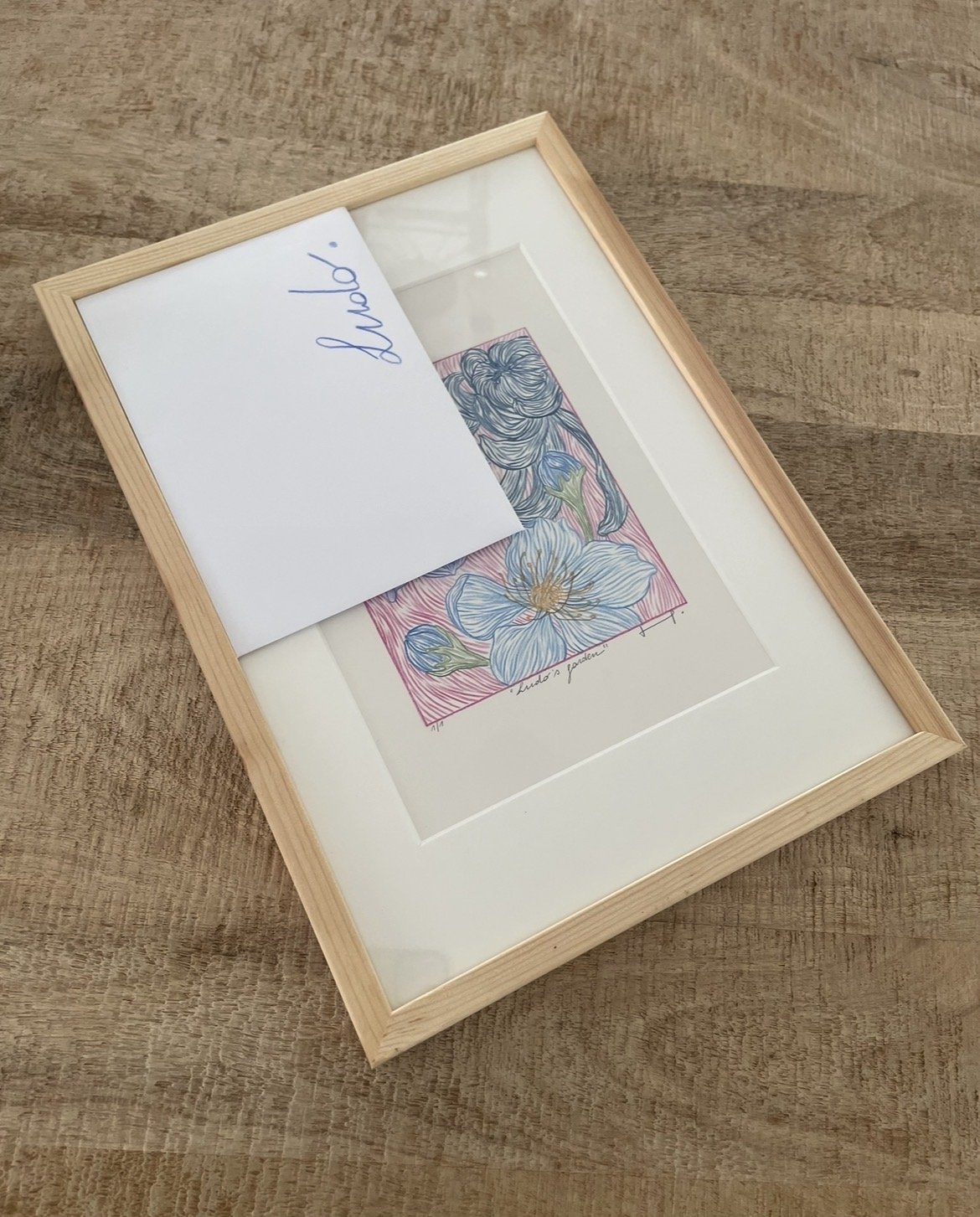 Framed botanical illustration of flowers on a wooden surface with a white card that says 'Hello!' placed on top.