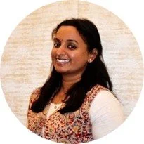 Portrait of a smiling woman with dark hair, wearing a patterned top and white long-sleeve shirt, against a light textured background.