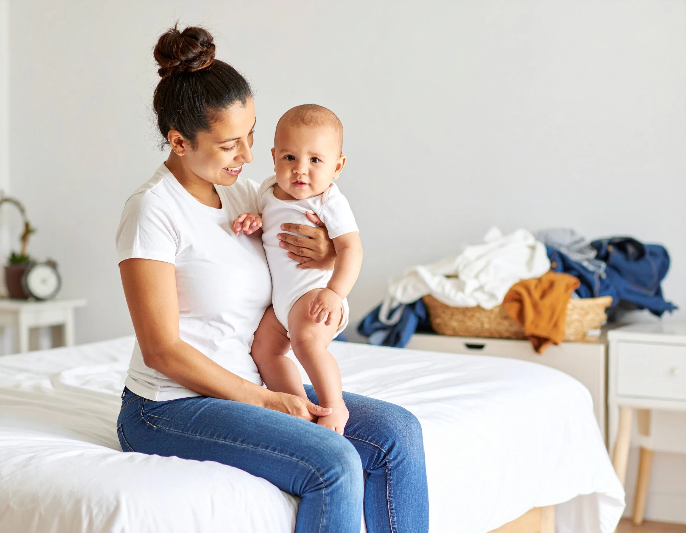 A woman (mom) with dark hair tied in a bun, wearing a white t-shirt and jeans, sitting on a bed and holding a baby in her arms. The baby, dressed in a white onesie, looks at the camera, and the woman is smiling. She loves taking care of her infant.