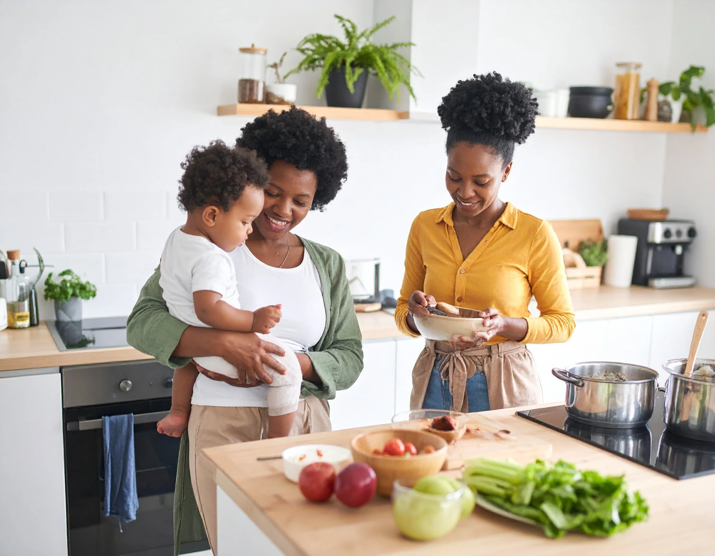 Two women (moms) and a small child in a modern kitchen preparing food together, with fresh vegetables on the counter.