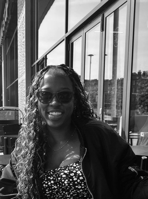 A woman with long curly hair wearing sunglasses smiling at the camera, sitting indoors with large windows and reflections of a cityscape behind her.