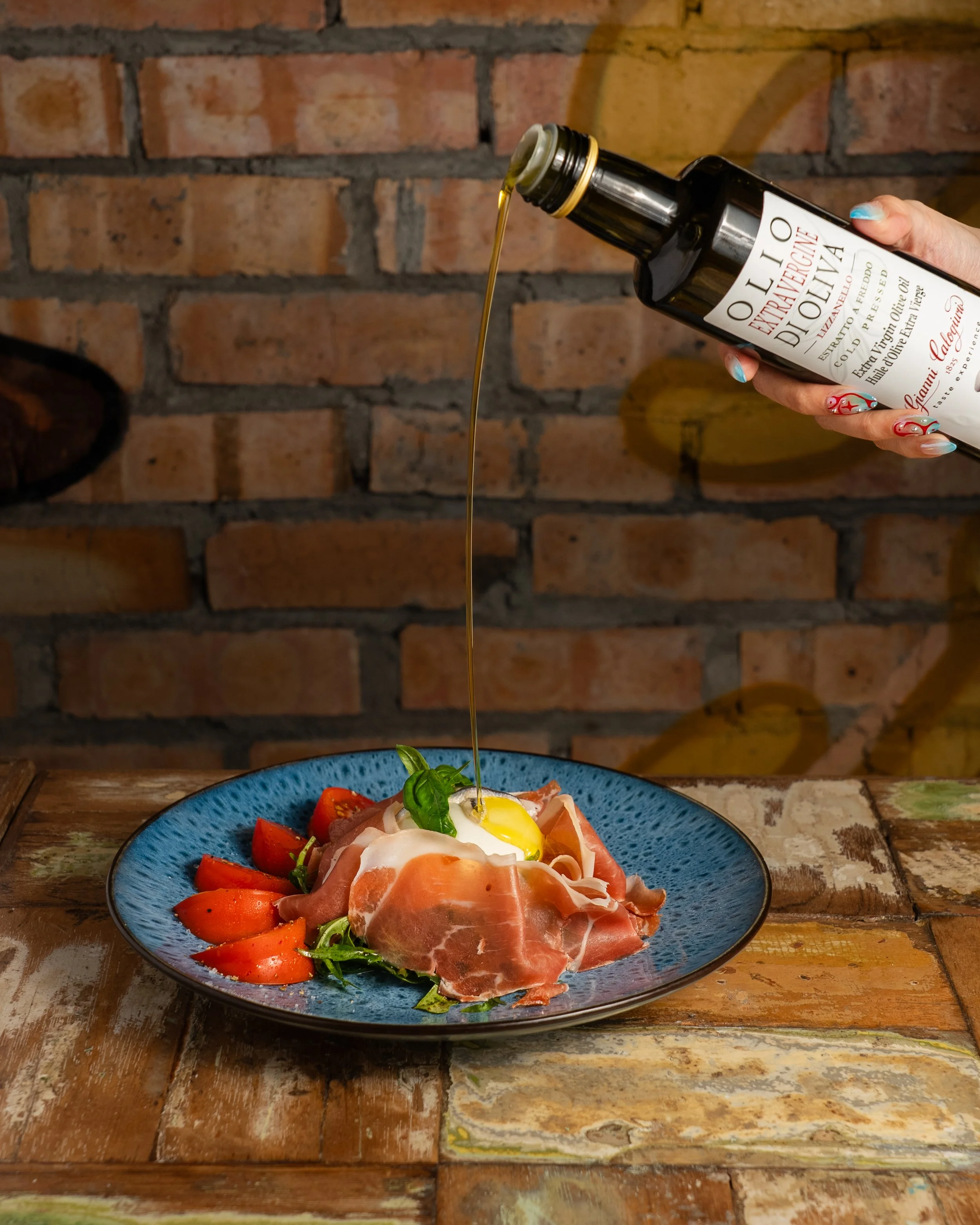 A plate of sliced tomatoes, prosciutto, and an over-easy egg, garnished with basil, on a blue dish. A hand is pouring olive oil from a dark bottle onto the dish, against a brick wall background.
