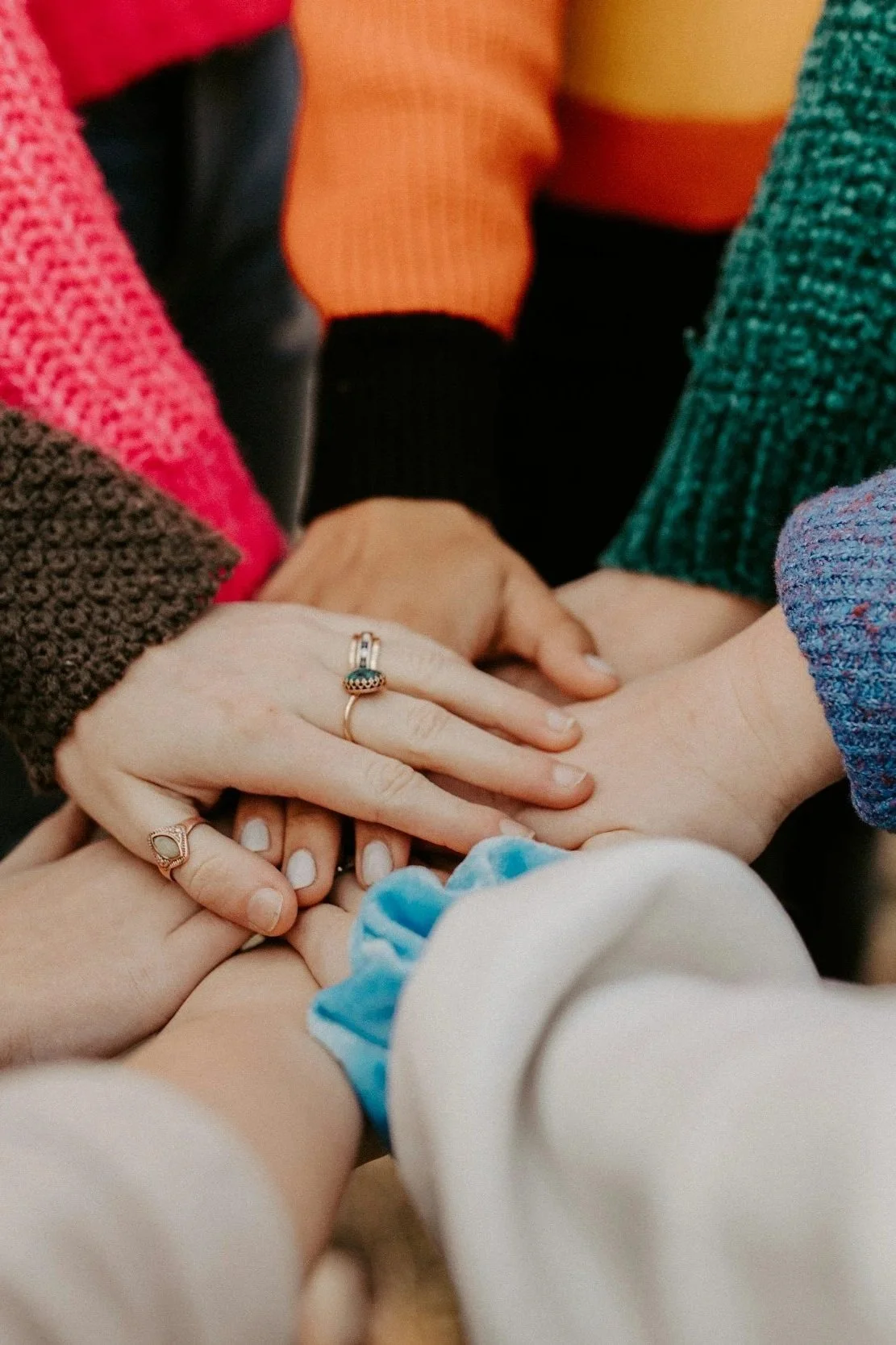 Multiple hands of diverse individuals stacked together, showing various rings and colorful sweaters, symbolizing unity and friendship.
