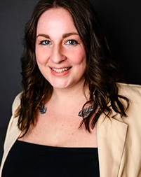 Portrait of a young woman with shoulder-length curly hair, smiling, wearing a black top and a beige blazer against a dark background.