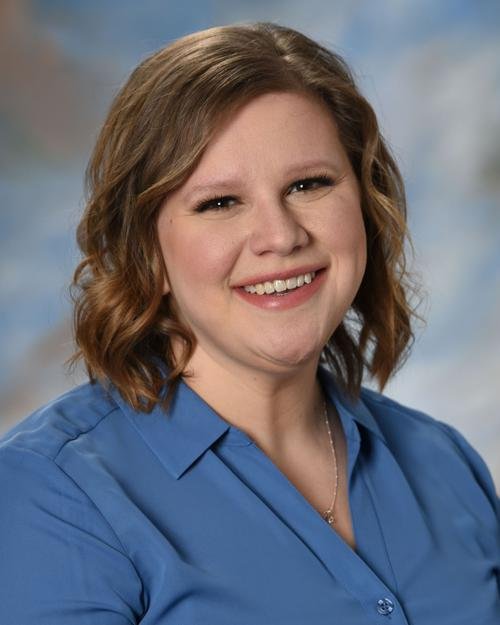 A smiling woman with shoulder-length wavy hair, wearing a blue collared shirt, against a blurred blue and beige background.