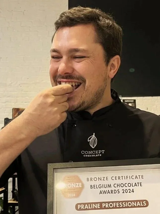 A man smiling and tasting a chocolate piece, holding a framed certificate from the Belgium Chocolate Awards 2024.