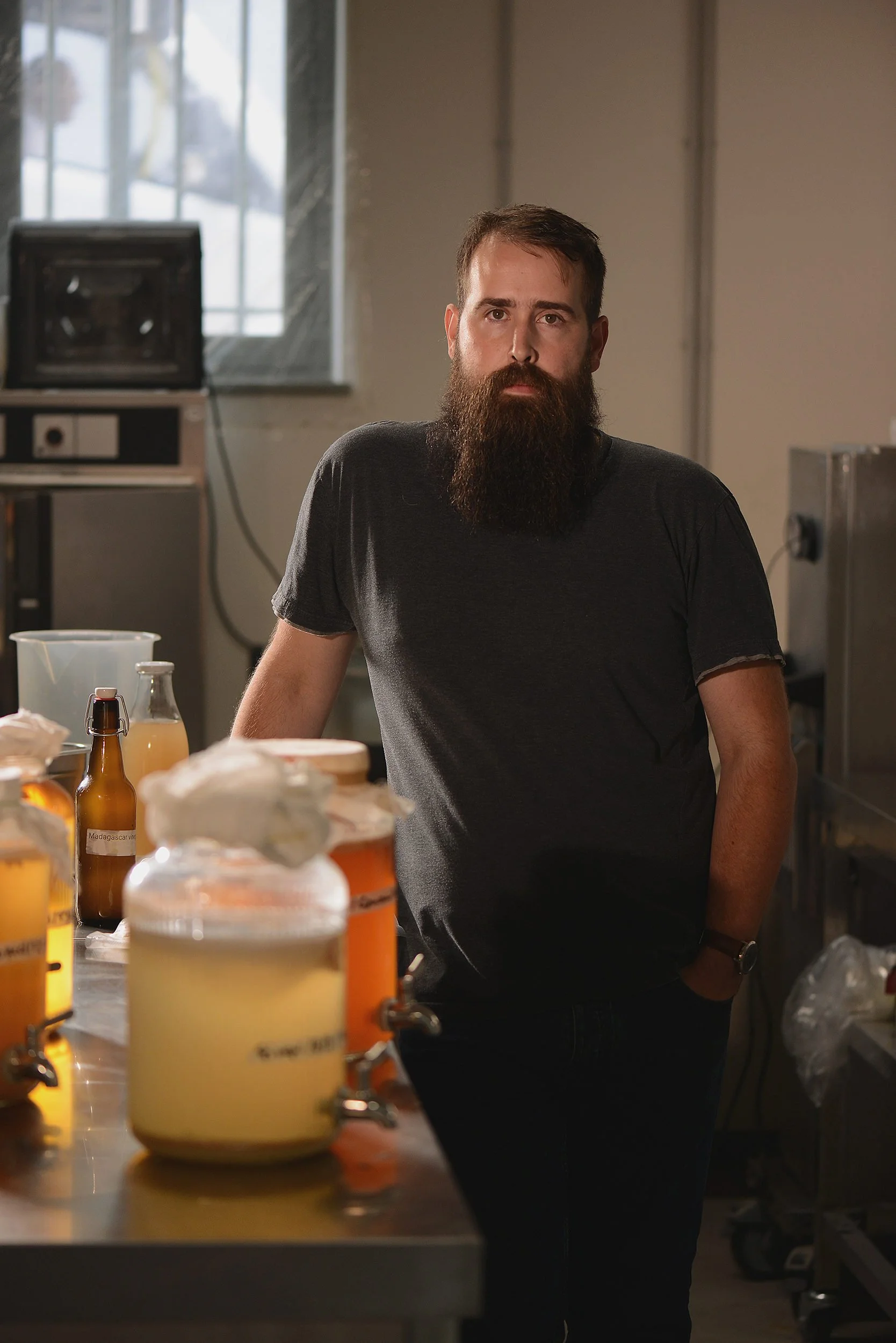 Man with a beard and black t-shirt standing in a kitchen or laboratory with jars and bottles on a table.