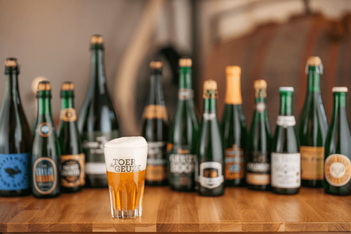 A beer glass with foam on top of the logo of Toer de Geuze, placed on a wooden table. Behind it are multiple bottles of different beers in a bar setting, with blurred background.