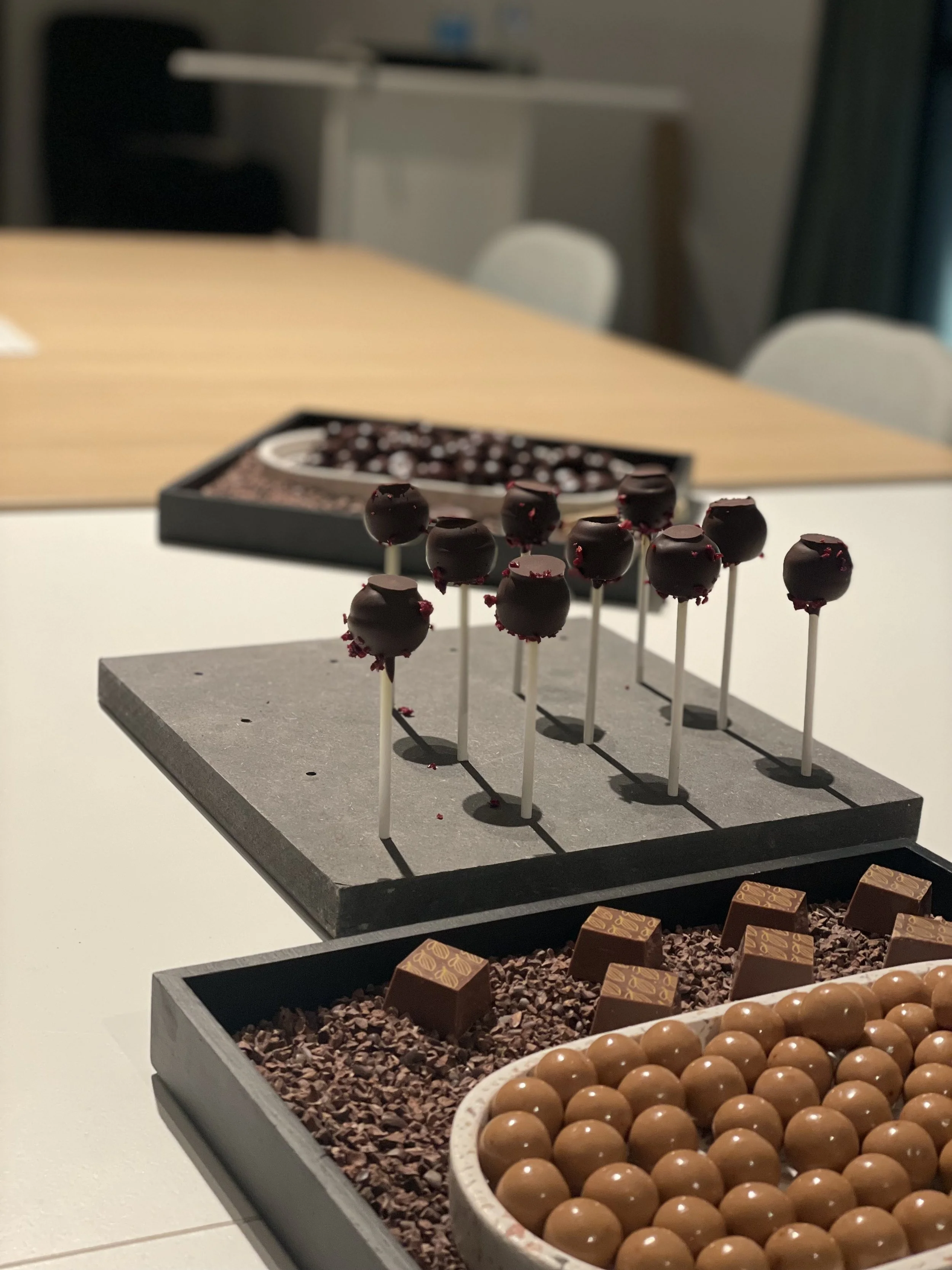 Assorted chocolates on display with chocolate-covered cake pops on sticks in the foreground, decorated with pink sprinkles, and a box of assorted chocolates in the background.