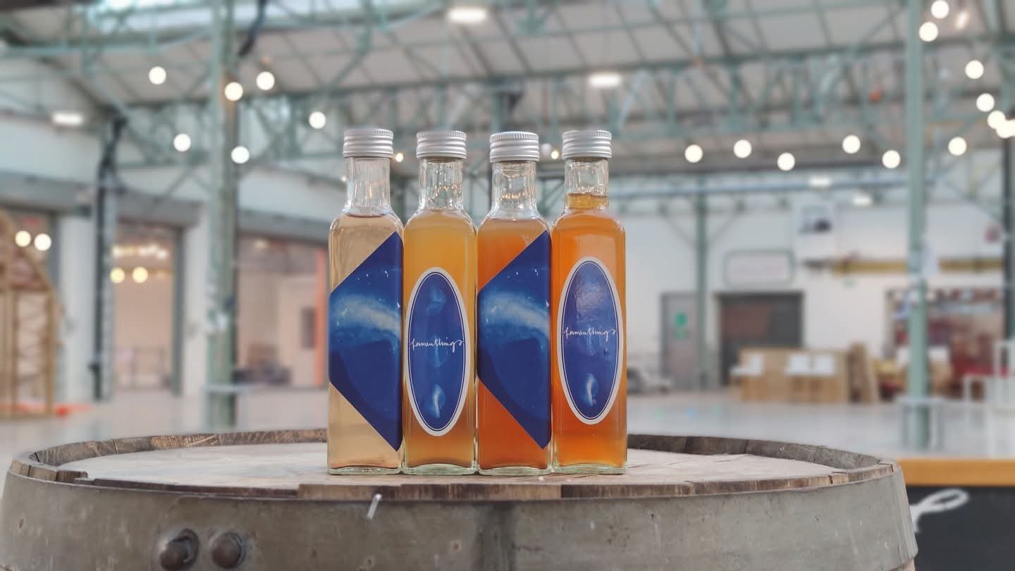 Four bottles with different colored liquids, placed on a wooden barrel in an indoor space with hanging lights and industrial architecture.