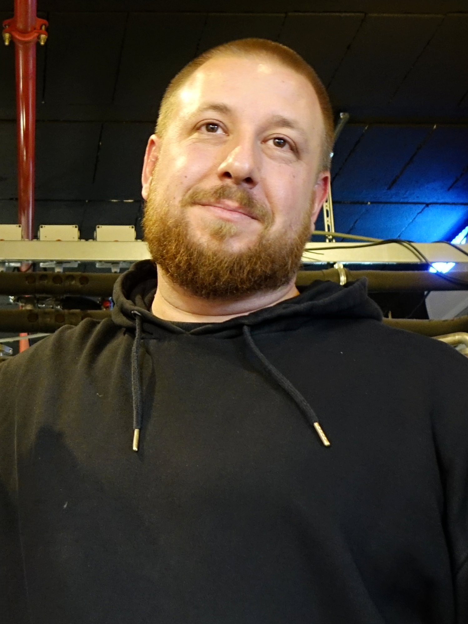 A man with a beard and short hair, wearing a black hoodie, smiling indoors with machinery and a dark ceiling in the background.