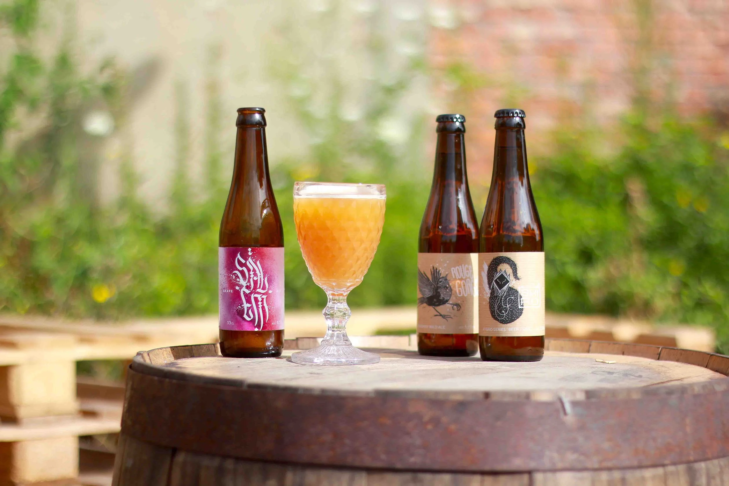 Three bottles of craft beer and a glass of beer with a textured surface, placed on a wooden barrel outdoors with a blurred green and reddish background.
