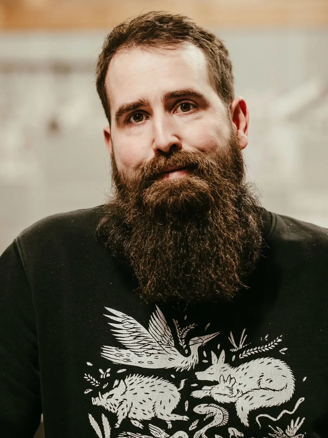 A man with a thick, full beard and short brown hair, wearing a black shirt with a white graphic of animals and plants.