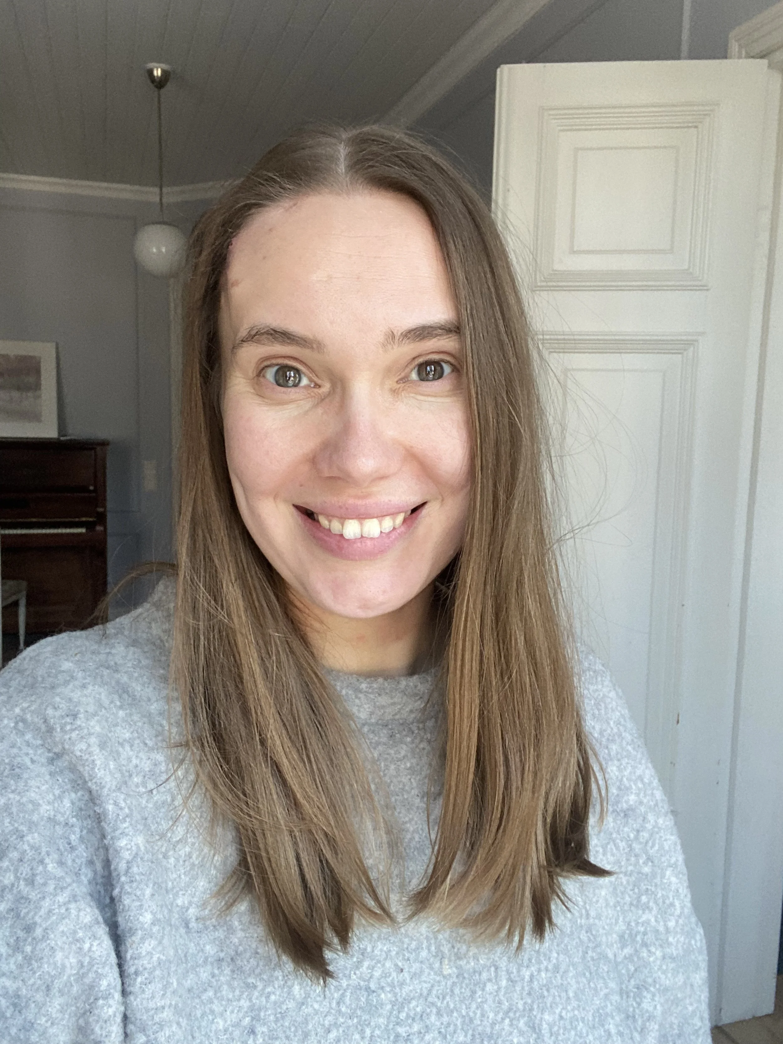 A woman with long brown hair smiling in a room with a light-colored wall, a white door, and a piano in the background.