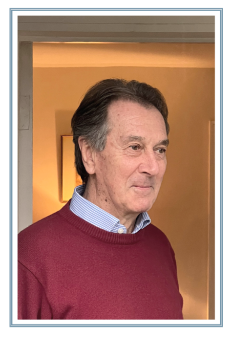 A middle-aged man with dark brown hair and light skin standing indoors, wearing a red sweater over a blue and white checkered shirt, looking slightly to the side with a slight smile.