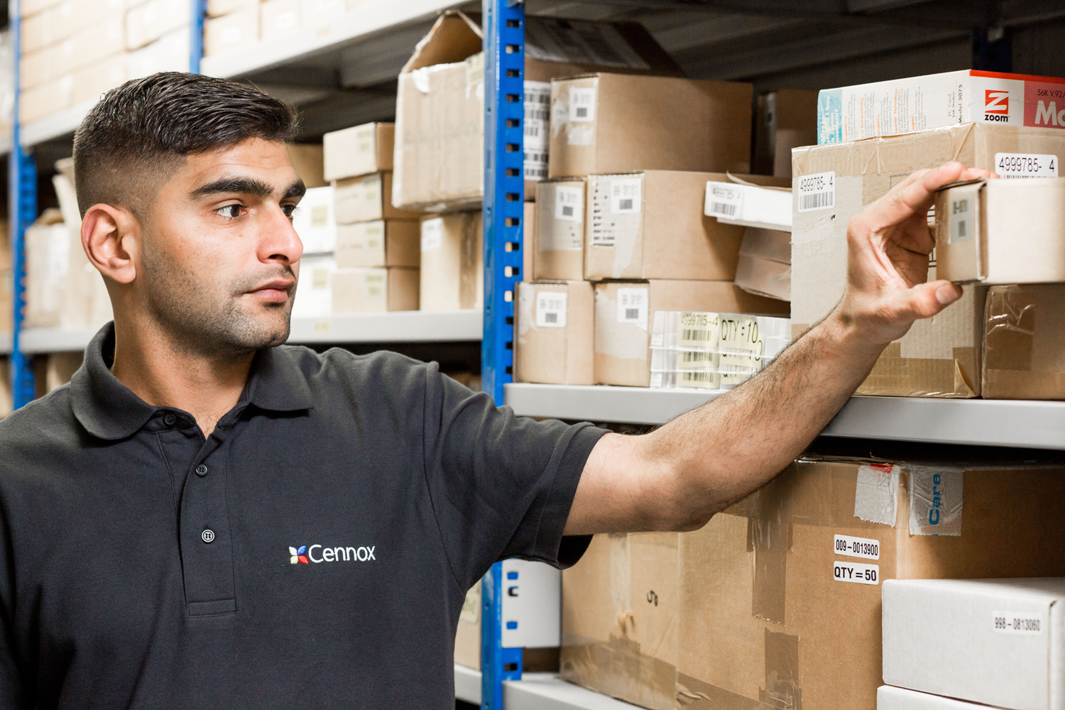 A man in a black polo shirt with the 'Cennox' logo is organizing or retrieving boxes from warehouse shelves filled with cardboard boxes.