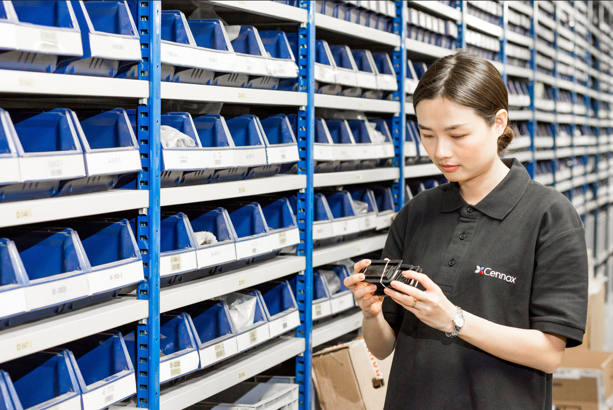 A woman wearing a black Cennox uniform inspecting items on a handheld device in a storage room with organized blue bins on metal shelves.