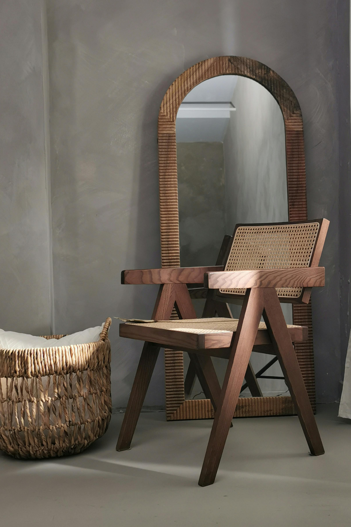 Un miroir en bois, un fauteuil en bois avec une assise en rotin, une corbeille en osier avec un coussin, dans une pièce avec un mur gris et un sol en béton.