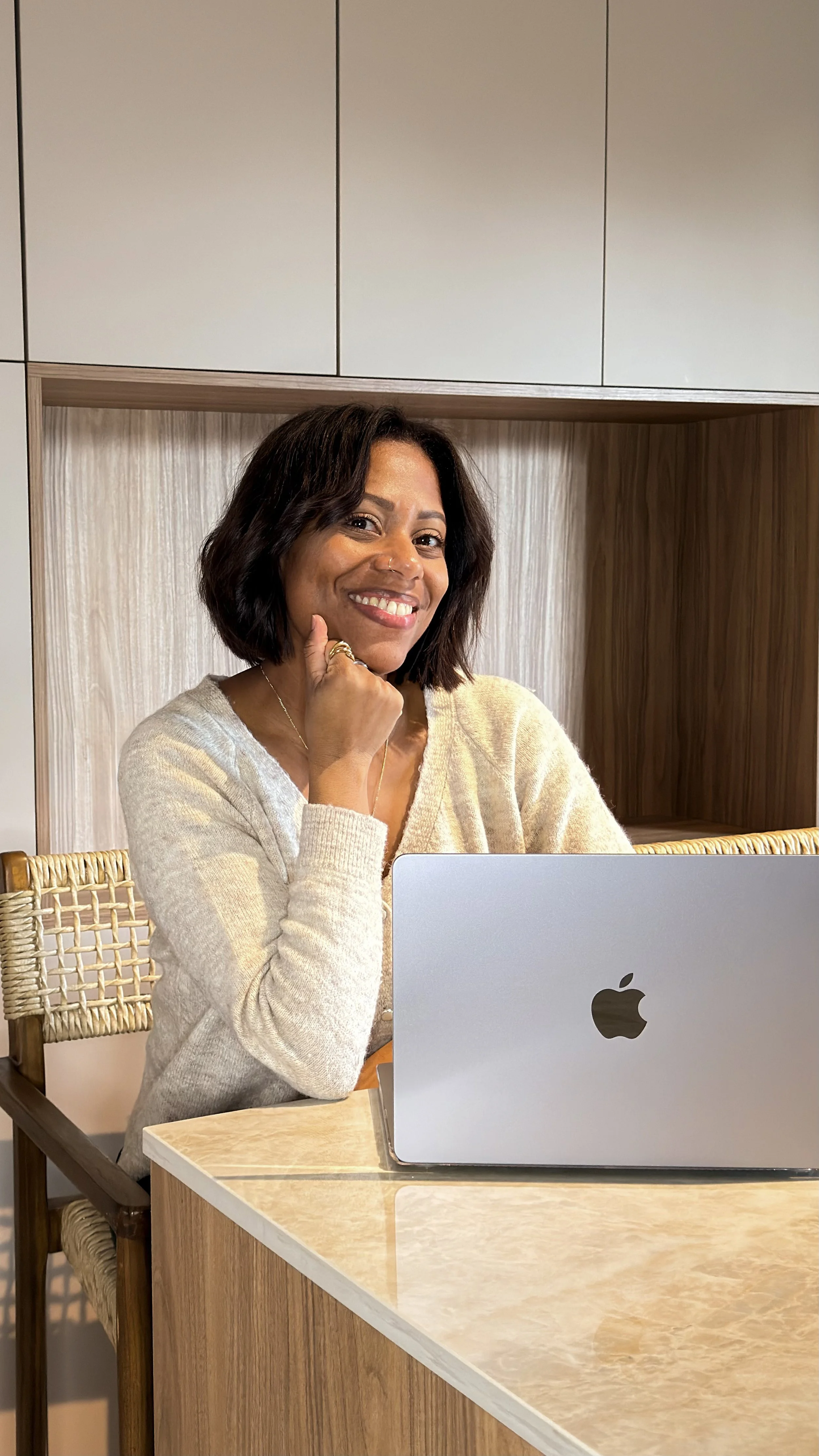Une femme souriante assise à une table avec un ordinateur portable Apple, portant un pull beige, dans un environnement intérieur moderne.