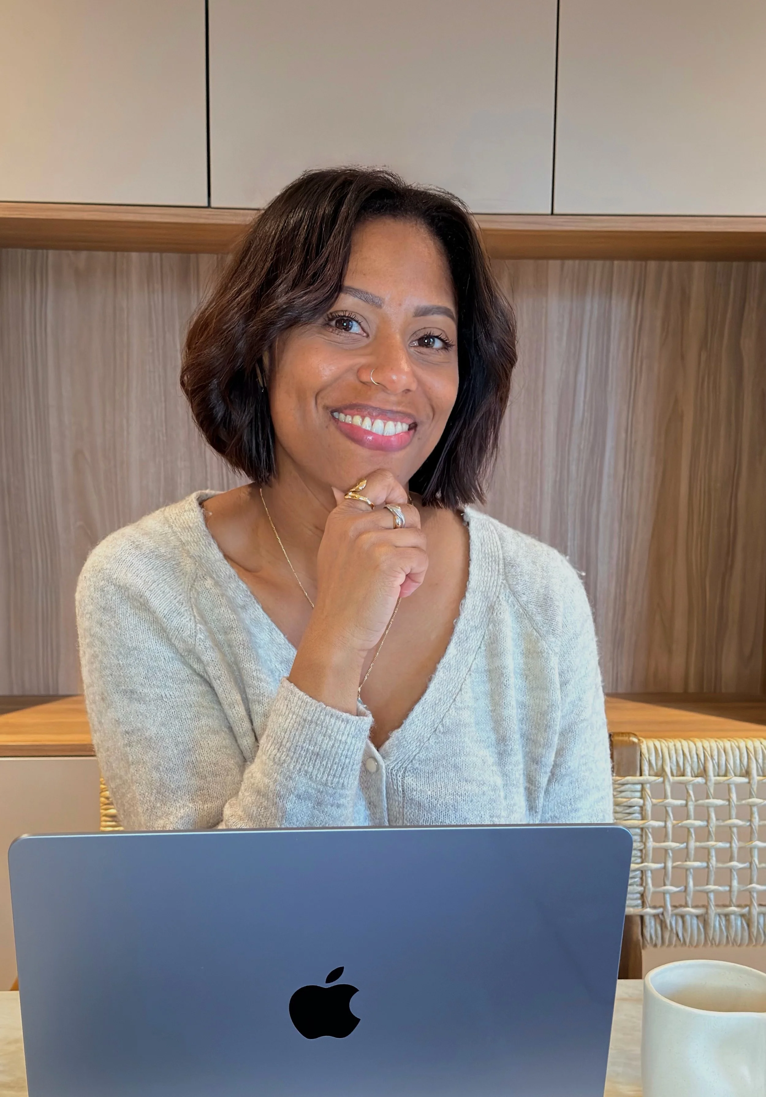 Une femme souriante assise devant un ordinateur portable Apple, dans un intérieur moderne avec des murs en bois. Elle porte un pull beige clair et a des bijoux en or.