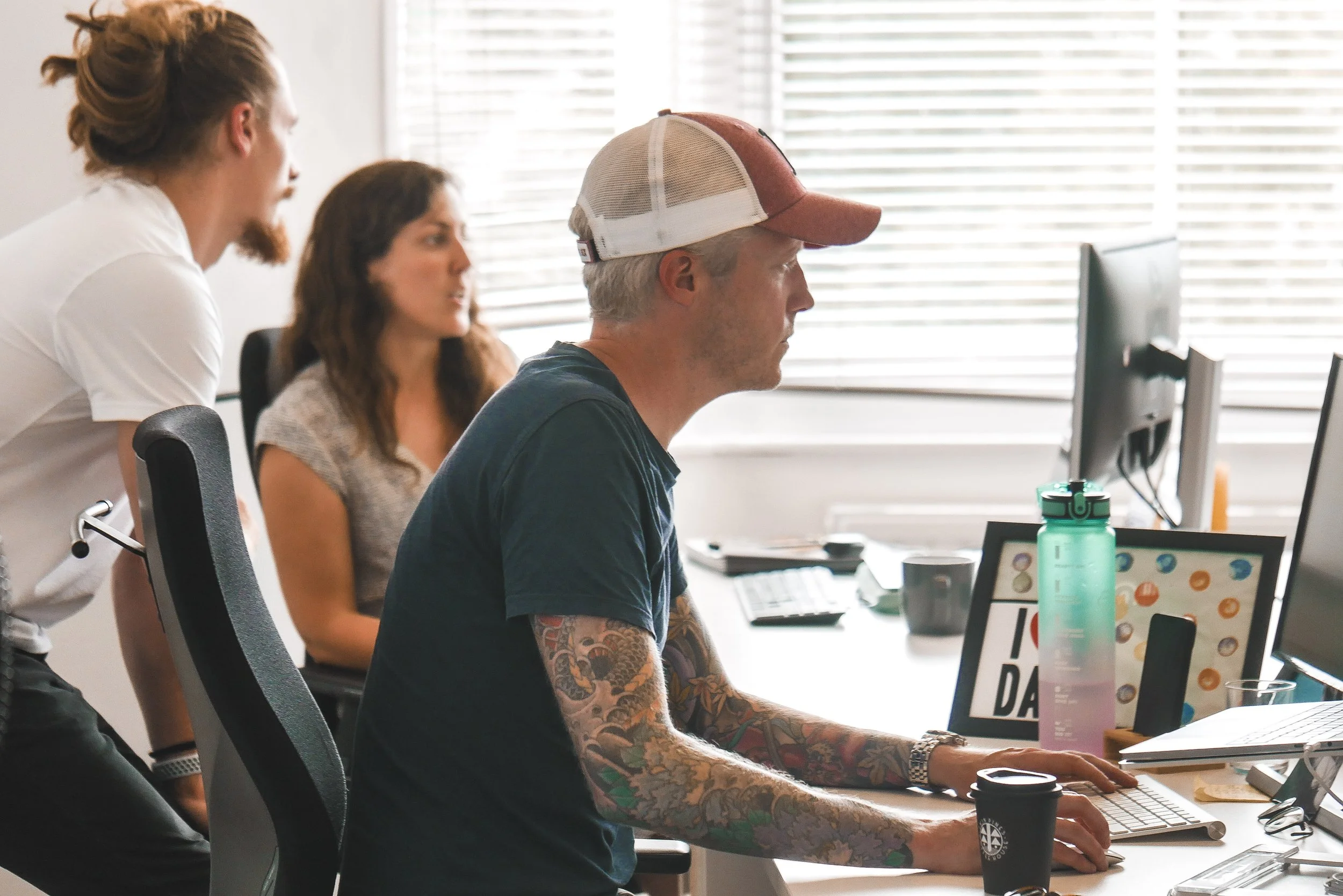 Three people working at desks in an office, with a man in a baseball cap and tattoos on his arm, and two women, one with brown hair and the other with red hair, looking at computers and monitors.