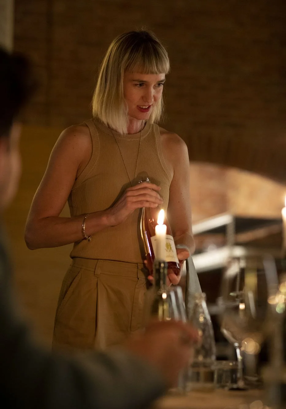 A woman with blonde hair in a sleeveless tan top holding a lit birthday candle in a bottle, celebrating with friends in a warmly lit restaurant or cafe.