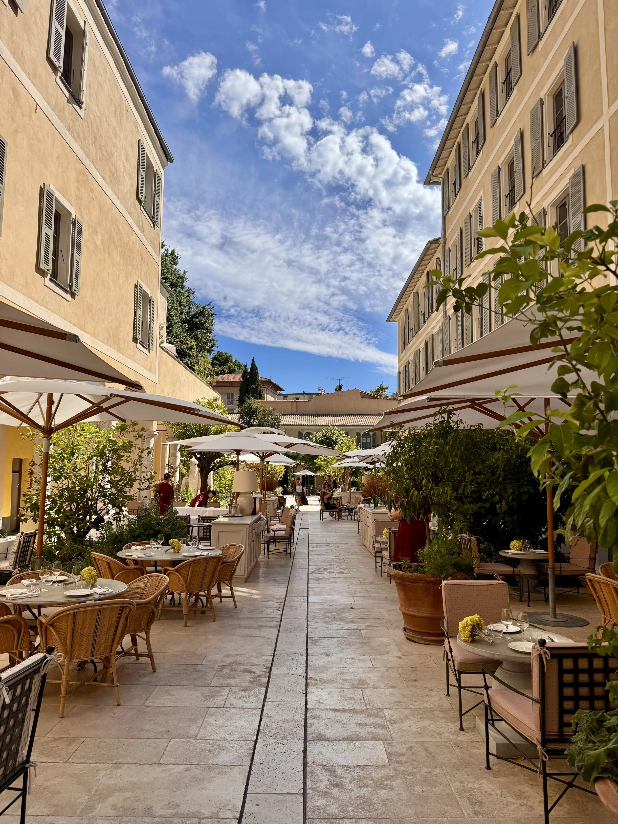 An outdoor restaurant or cafe patio with tables and chairs, some with yellow flowers, shaded by large white umbrellas. Tall potted plants and trees line the sides. Two large buildings with multiple windows and shutters flank the patio under a partly cloudy blue sky.