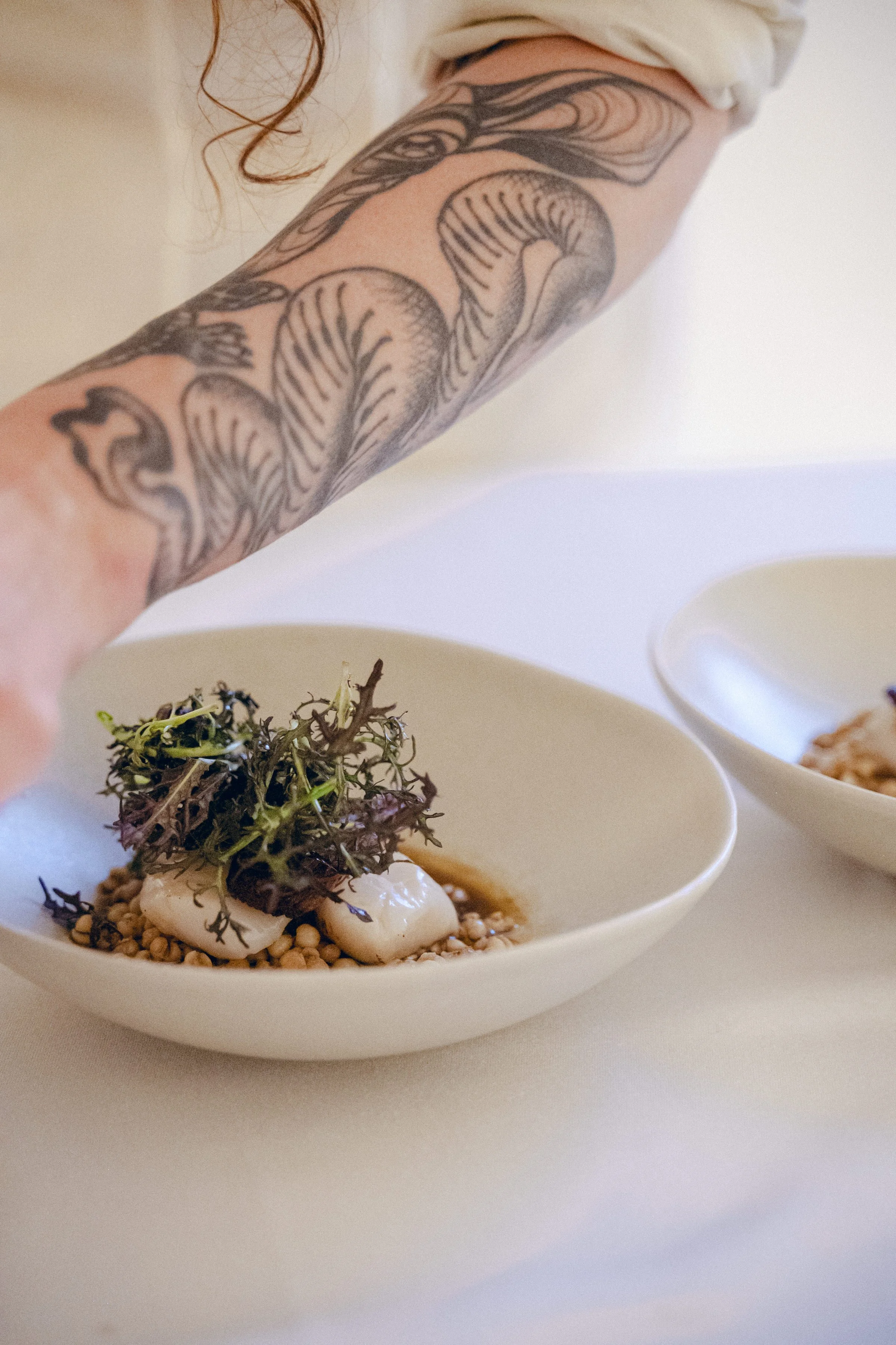 Close-up of a tattooed arm placing a plated dish with microgreens, seafood, and a sauce in a white bowl on a white surface.