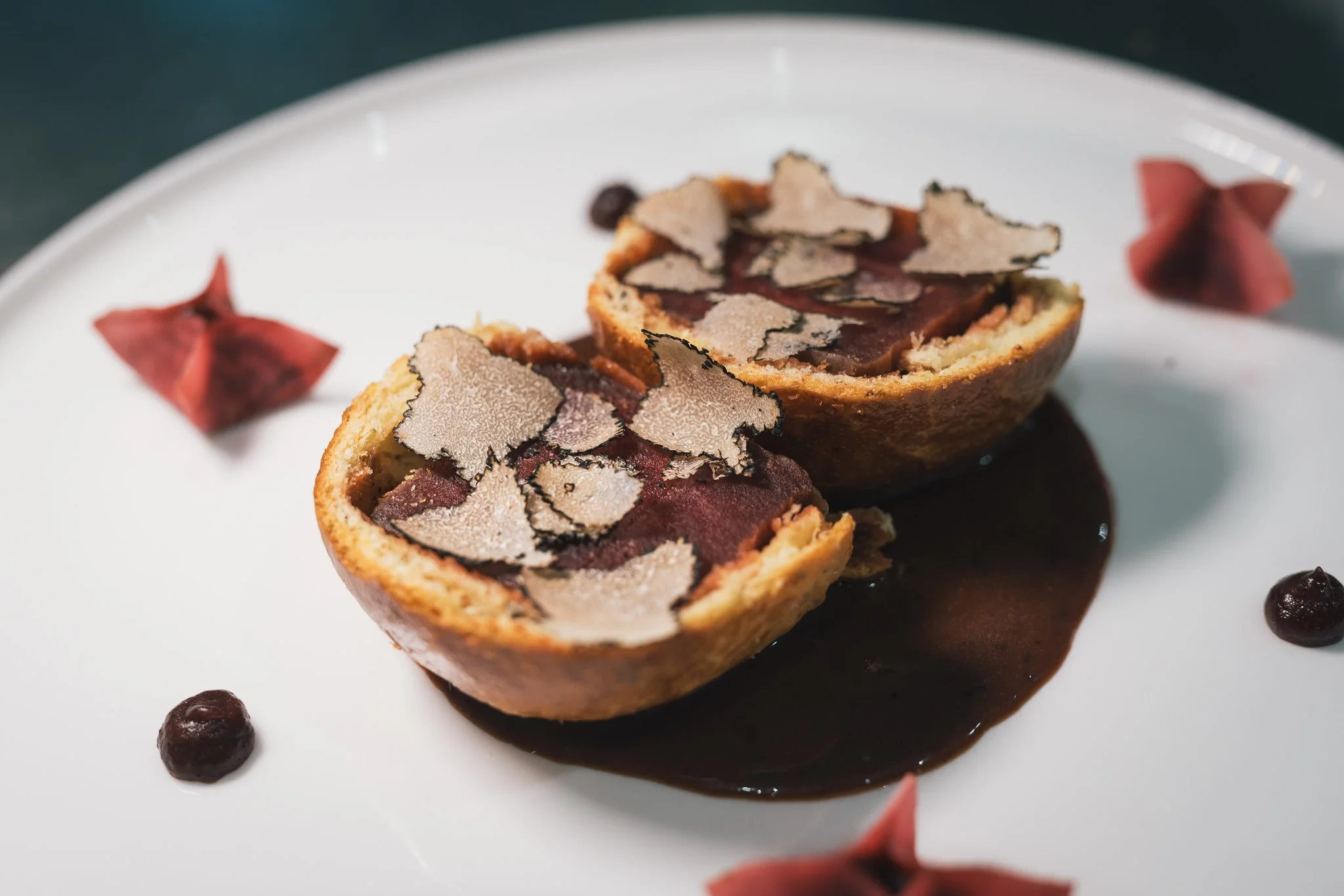 A plated dessert featuring a halved cherry tart topped with sliced truffle on a white plate with decorative pink and purple dollops and a dark sauce.