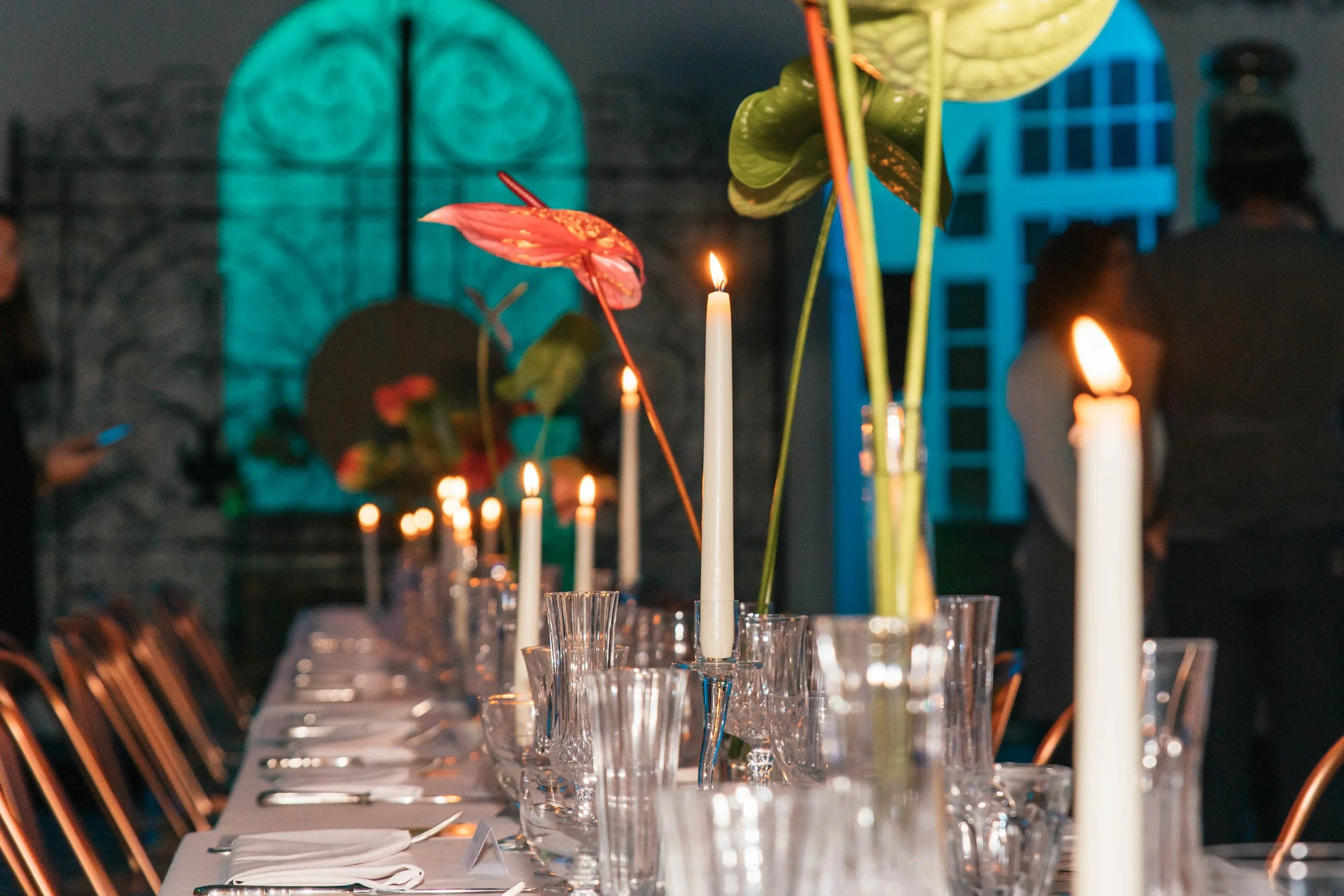 Elegant dining table set with tall white candles, glassware, and colorful floral centerpieces in a dimly lit room with blue and teal lighting.