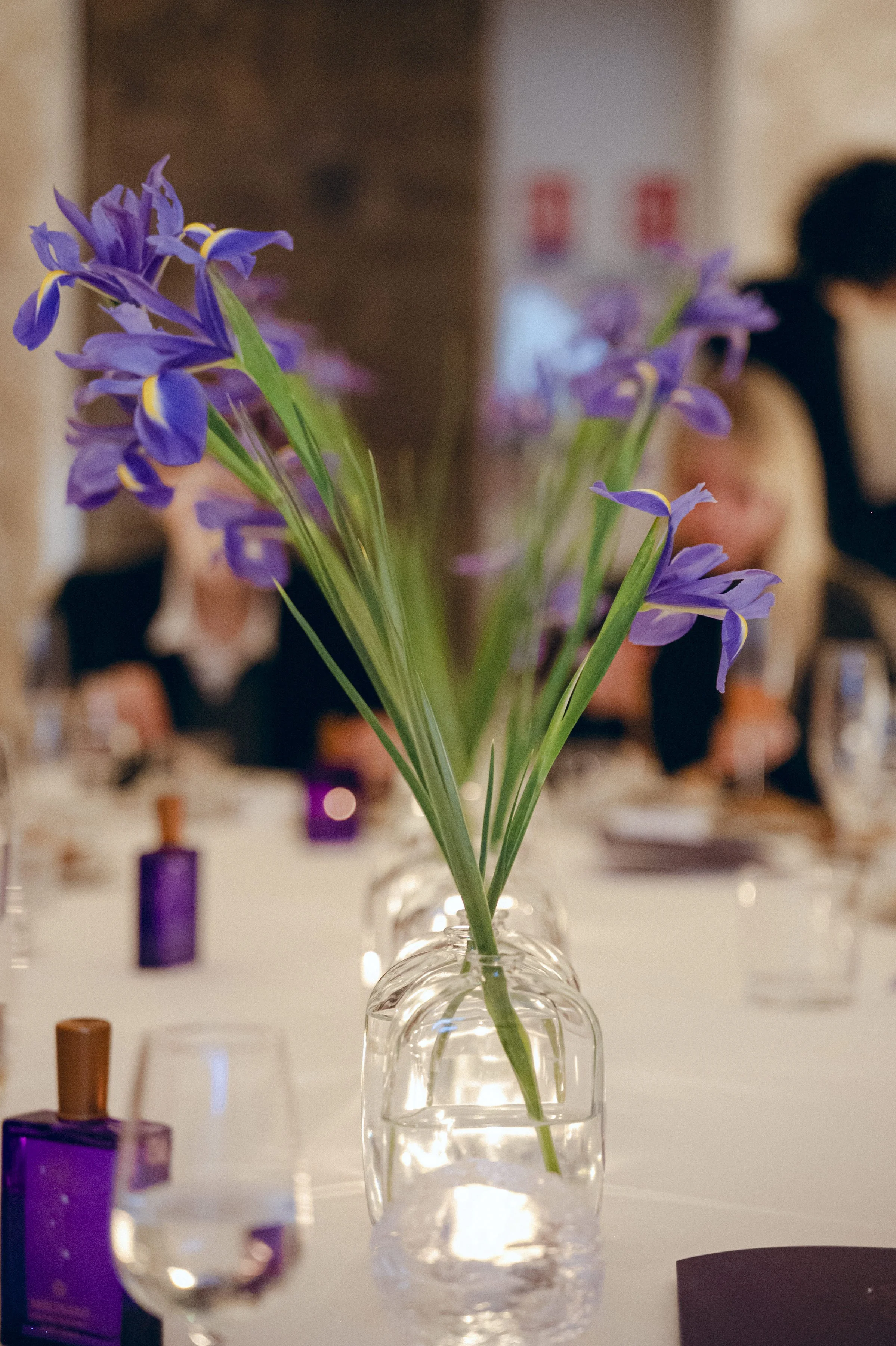 Purple flowers in a clear glass vase on a table at a gathering event.