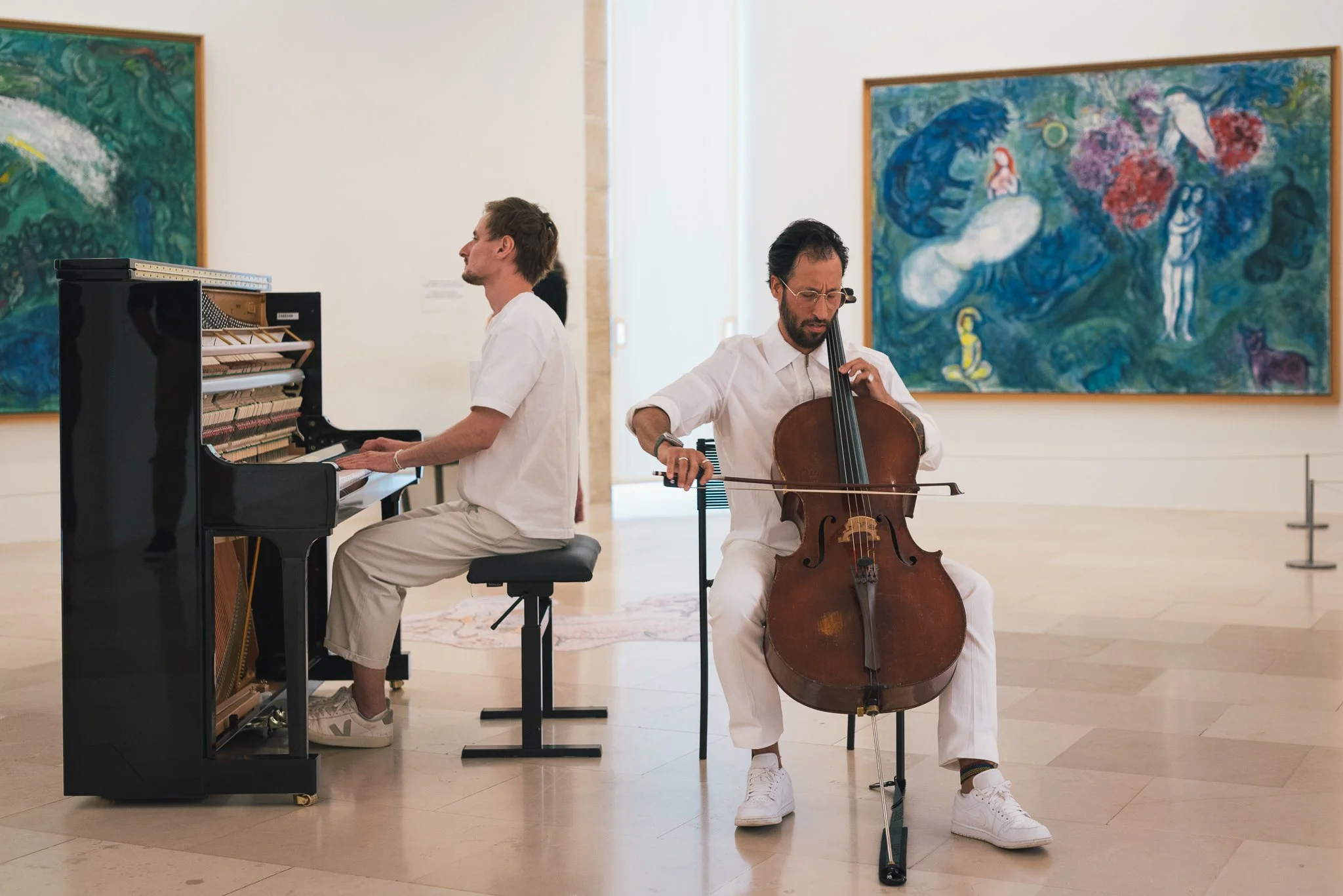 Two men in white clothing playing musical instruments in an art gallery. The man on the left is playing a black grand piano, and the man on the right is playing a cello. There are colorful paintings on the walls behind them.