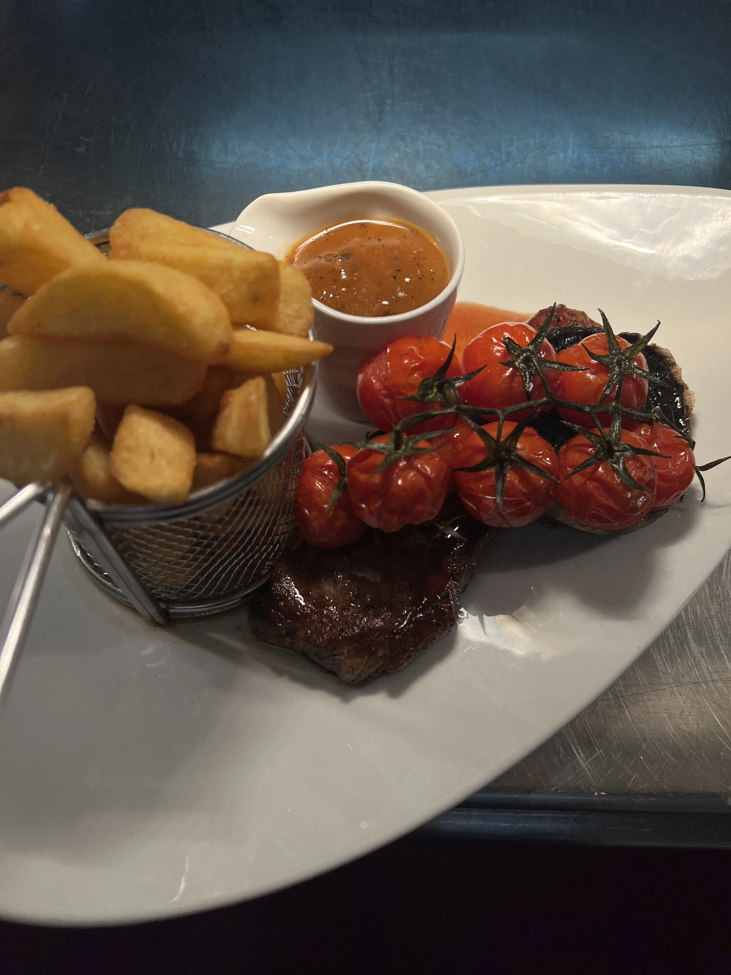 A plate of food with french fries, grilled tomatoes on the vine, a piece of cooked beef, and a small bowl of dipping sauce.