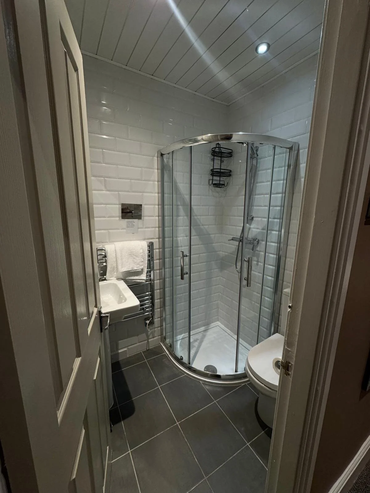 Small bathroom with a corner shower with glass doors, white tiled walls, a small sink with a towel, a floating toilet, and black floor tiles.
