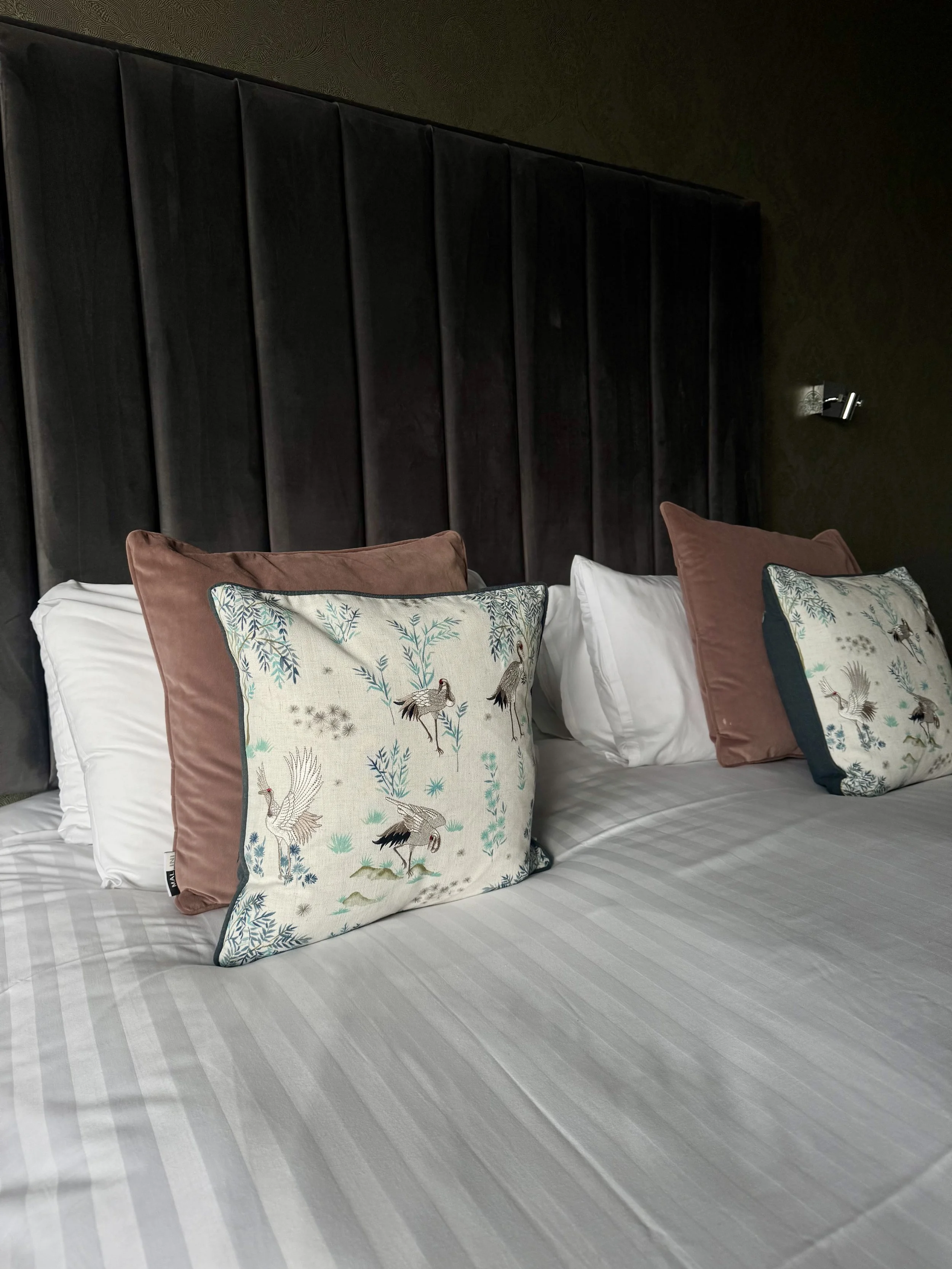 A hotel bed with decorative pillows and a padded black headboard wall.