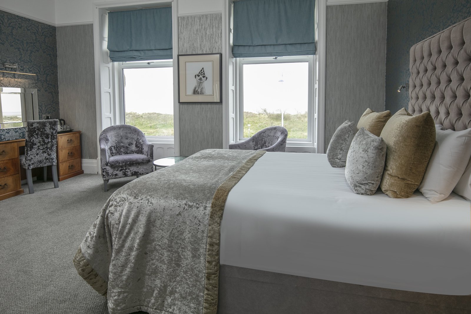 Hotel room with a large bed, multiple pillows, two armchairs, a small round table, a wooden desk, and two windows with blue curtains overlooking a grassy landscape.