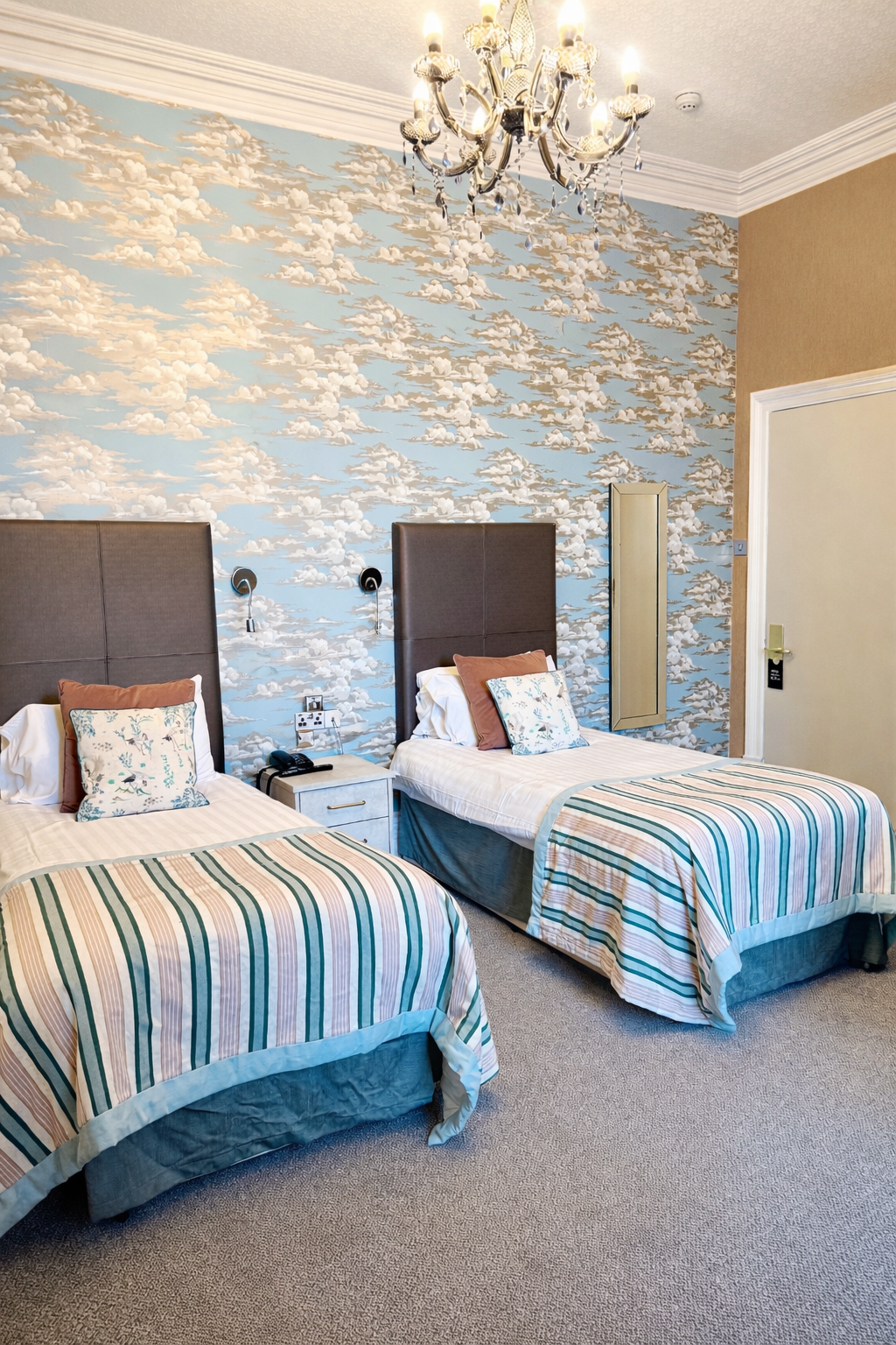 Hotel room with two beds, striped bedding, floral pillows, cloud-themed wallpaper, chandelier ceiling light, and bedside table with telephone