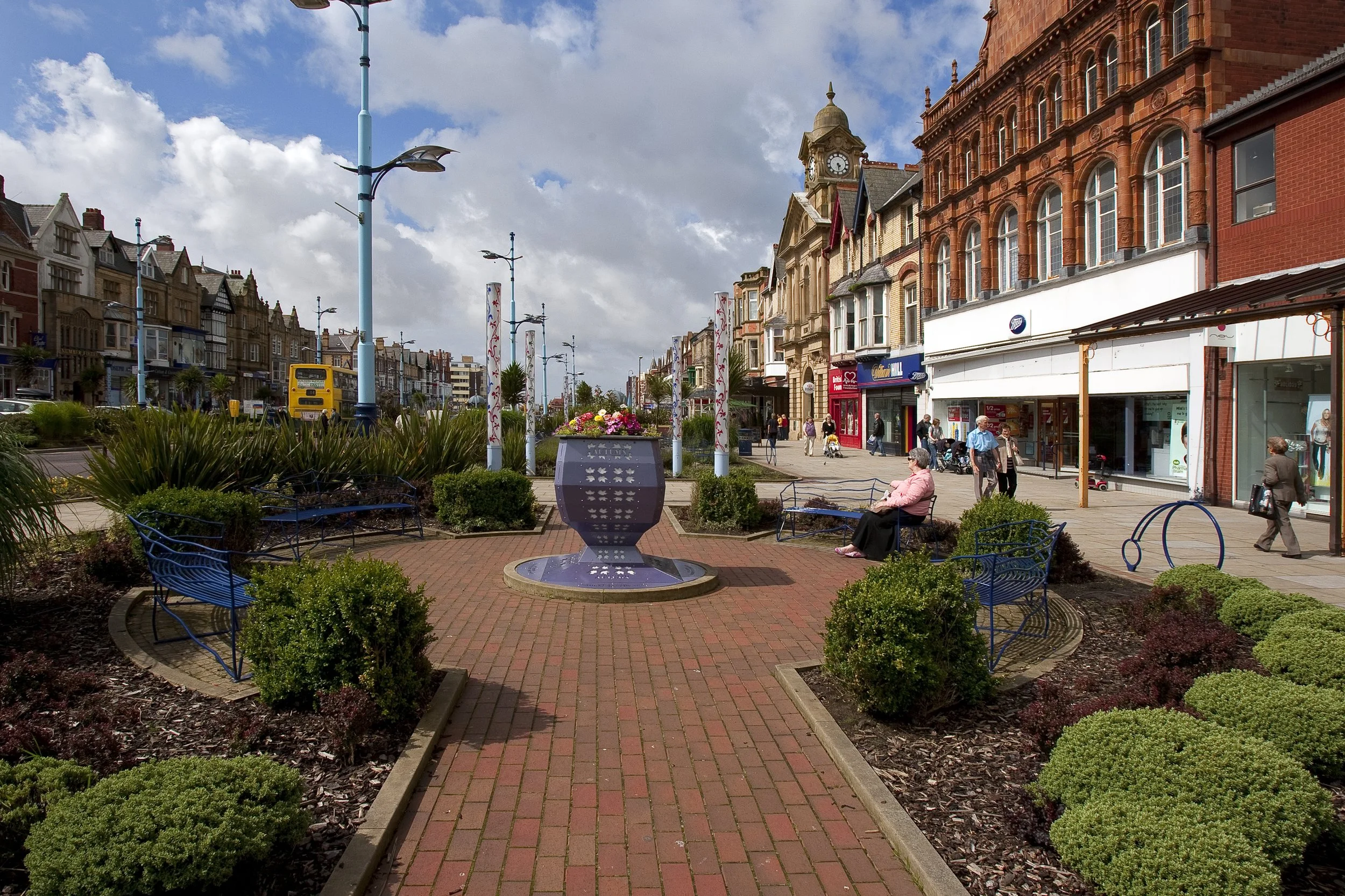A lively street scene with shops, pedestrians, and a central decorative fountain with flowers, surrounded by benches and landscaped bushes, under a partly cloudy sky.
