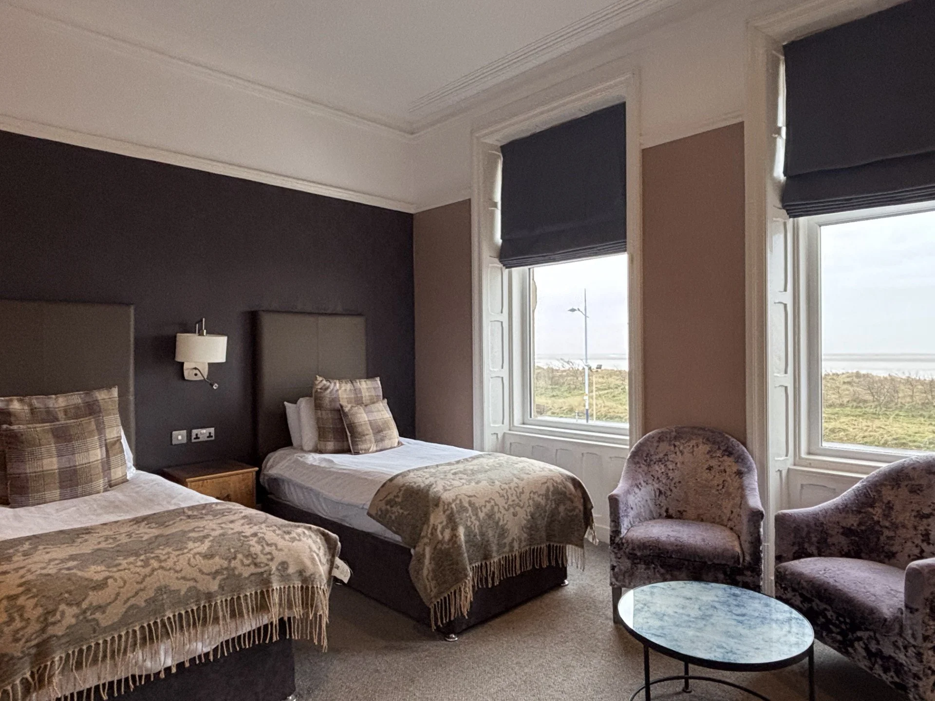 A hotel room with two single beds, plaid pillows, gray headboards, wall-mounted lamps, a nightstand, two large windows with black Roman shades, and a seating area with two velvet armchairs and a round marble-topped table.