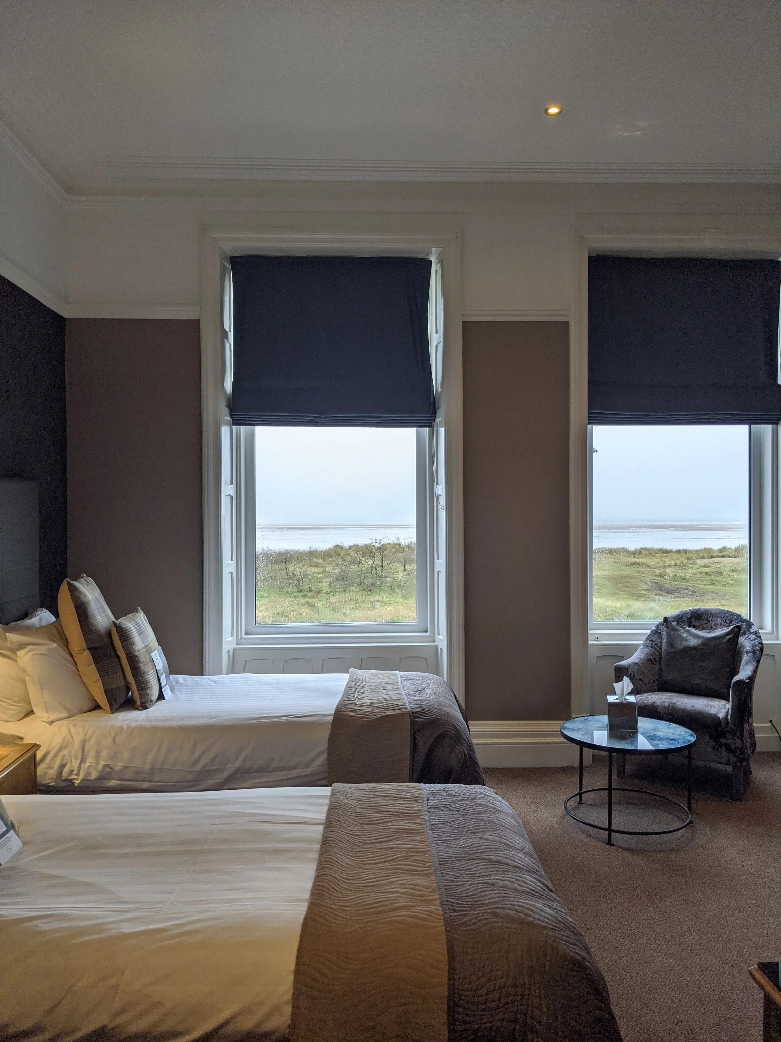 A hotel room with two beds, dark blue window shades, and a view of a grassy landscape and water outside. There is a side table with a tissue box and an armchair next to a small round table.